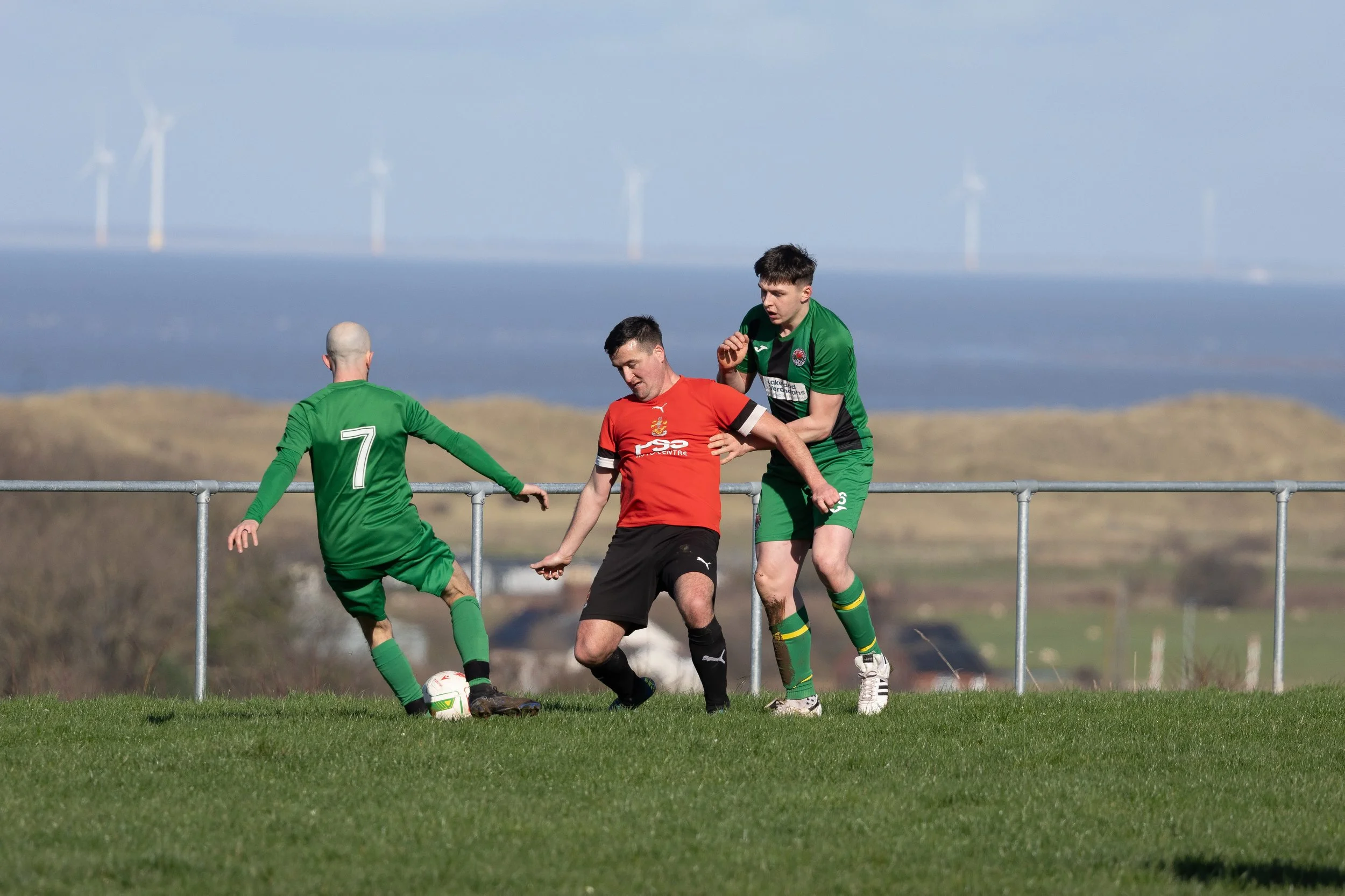 Two soccer players in green jerseys and one player in a red jersey competing for the ball on an outdoor field, with wind turbines in the background.