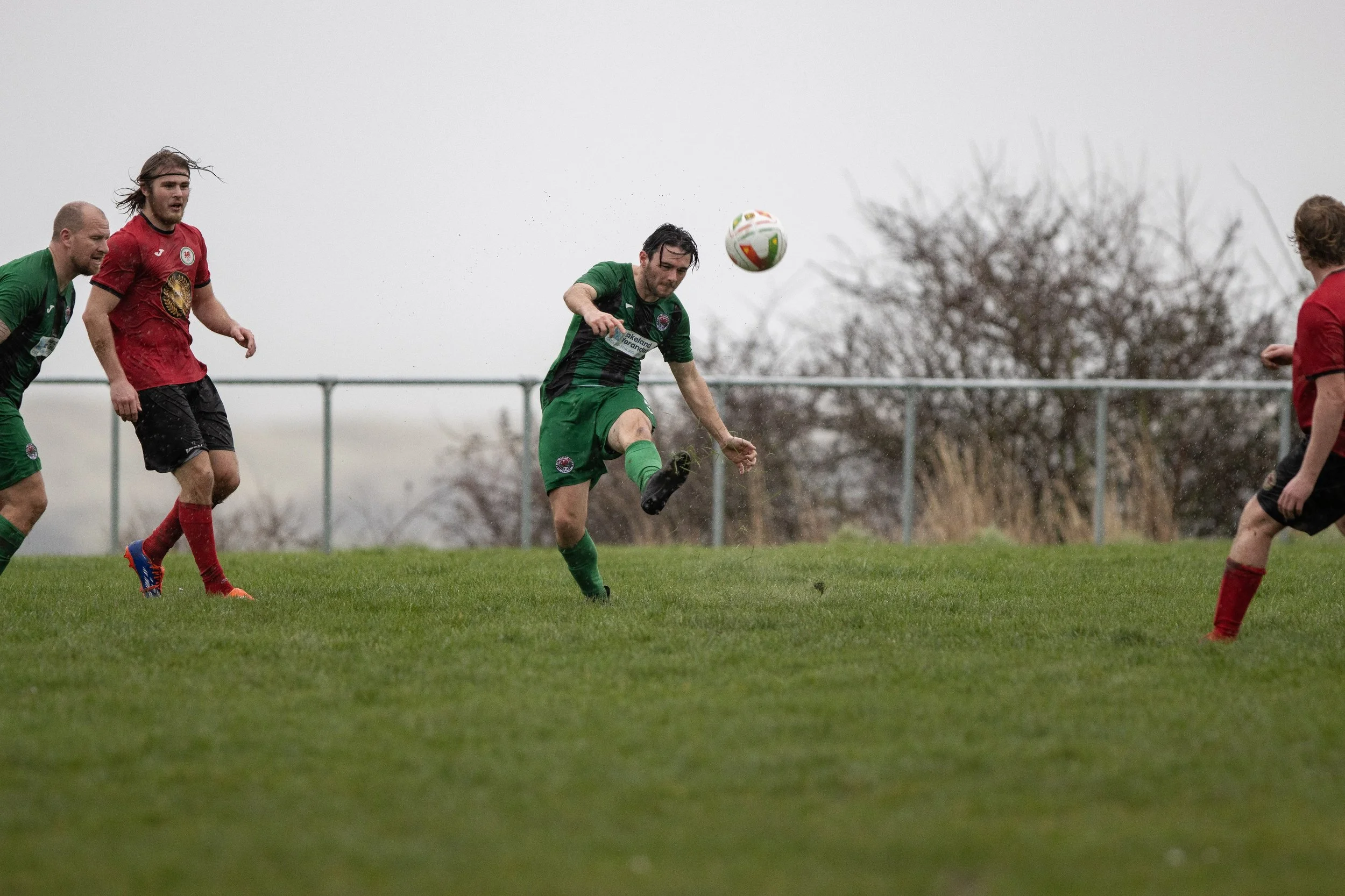 Soccer players in action on a grassy field during a match, with a cloudy sky and bare trees in the background.