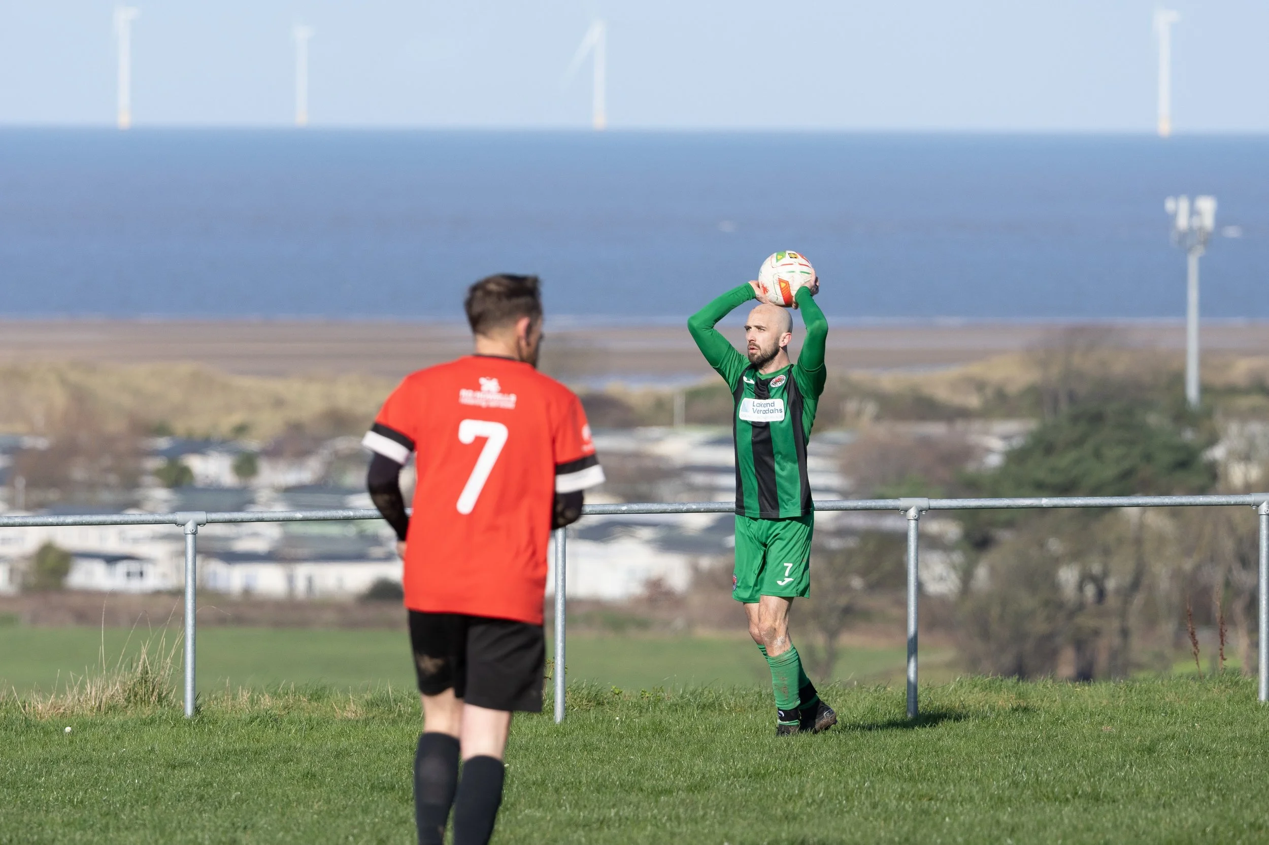 A soccer player in a green uniform holding a ball above his head while standing near a fence, with another player in a red jersey numbered 7 facing him on a grassy field, with wind turbines and a body of water in the background.
