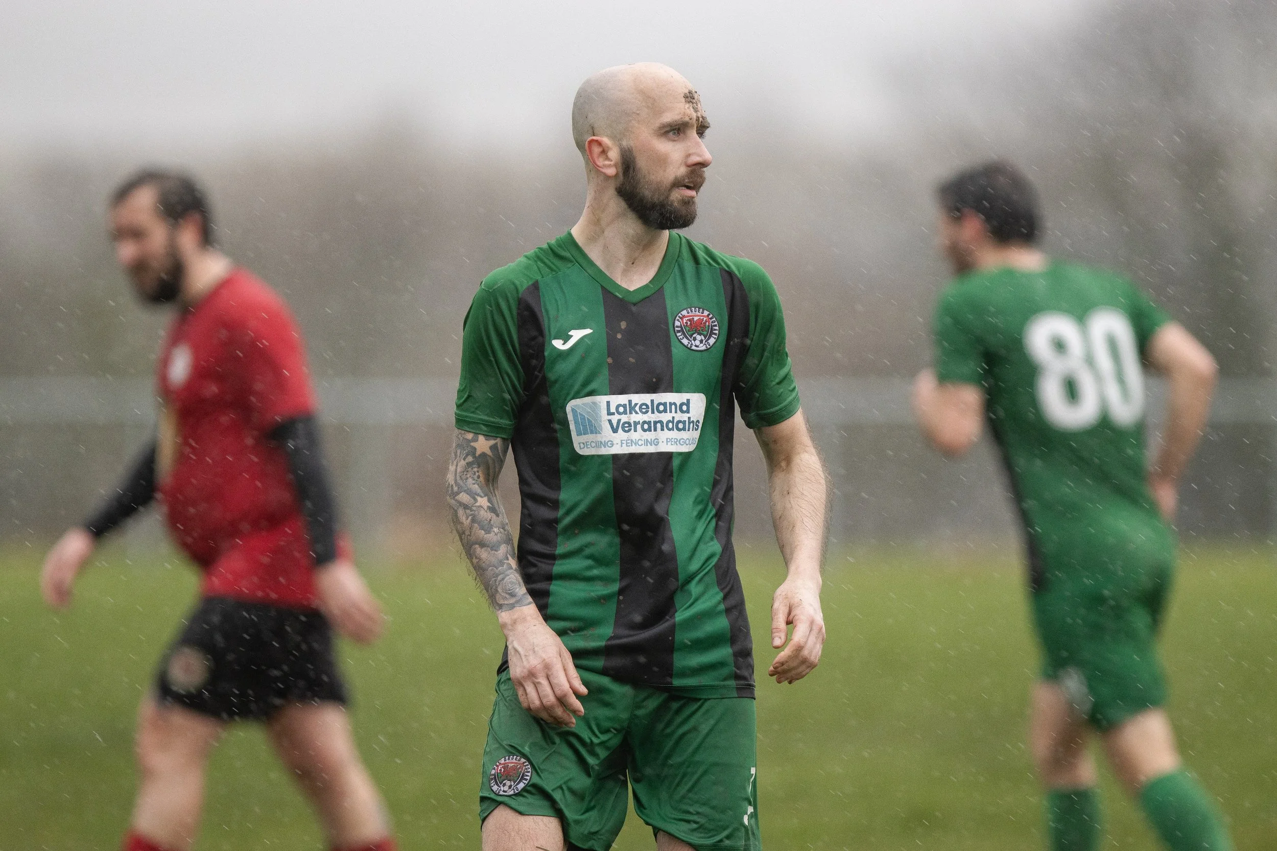 A soccer player in a green and black jersey with tattoos on his right arm stands on the field during a game in the rain, with other players blurred in the background.
