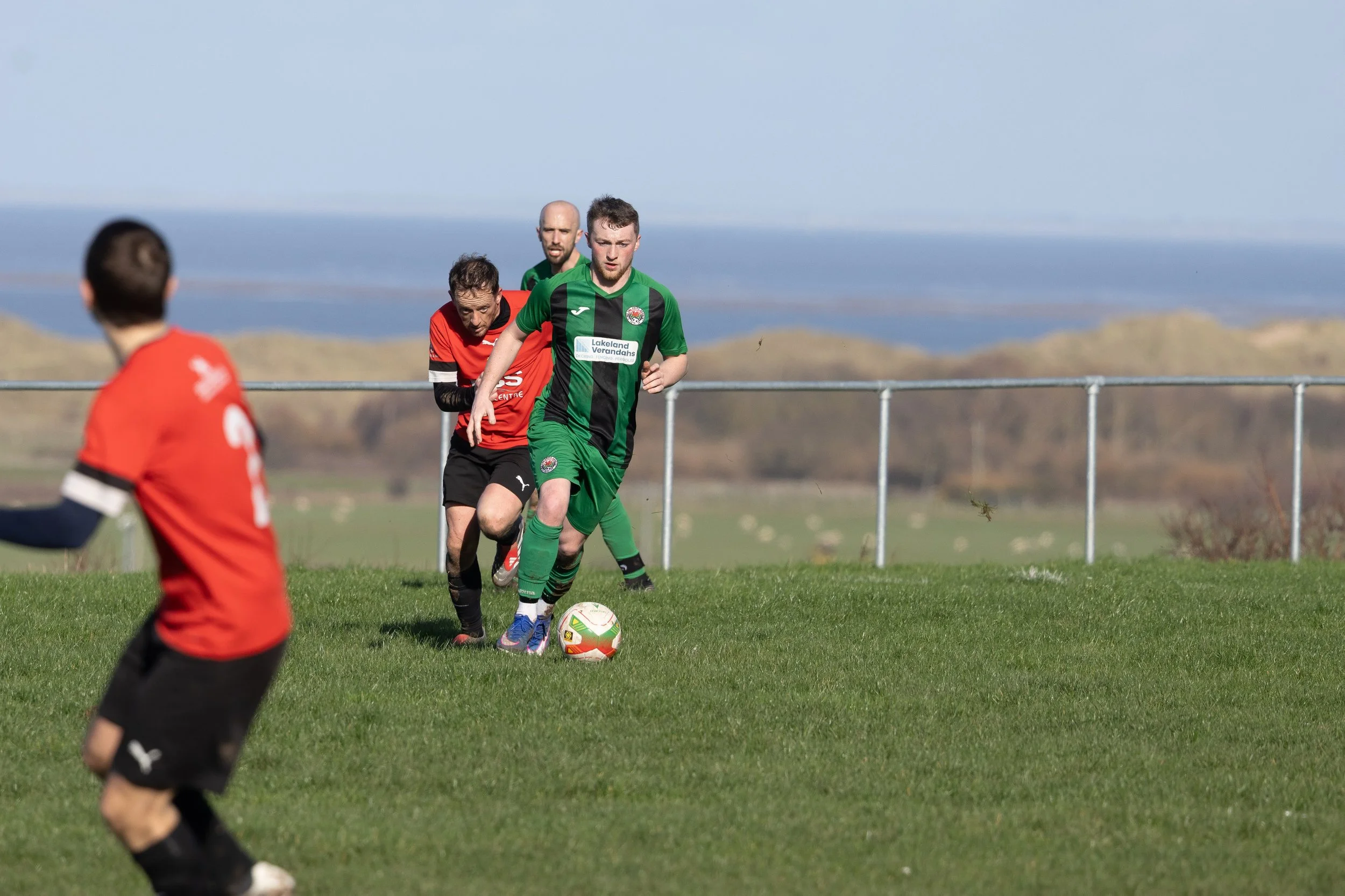 Soccer players in action, with one in a green and black uniform dribbling the ball while others in red and black uniforms pursue during a match on a grassy field.