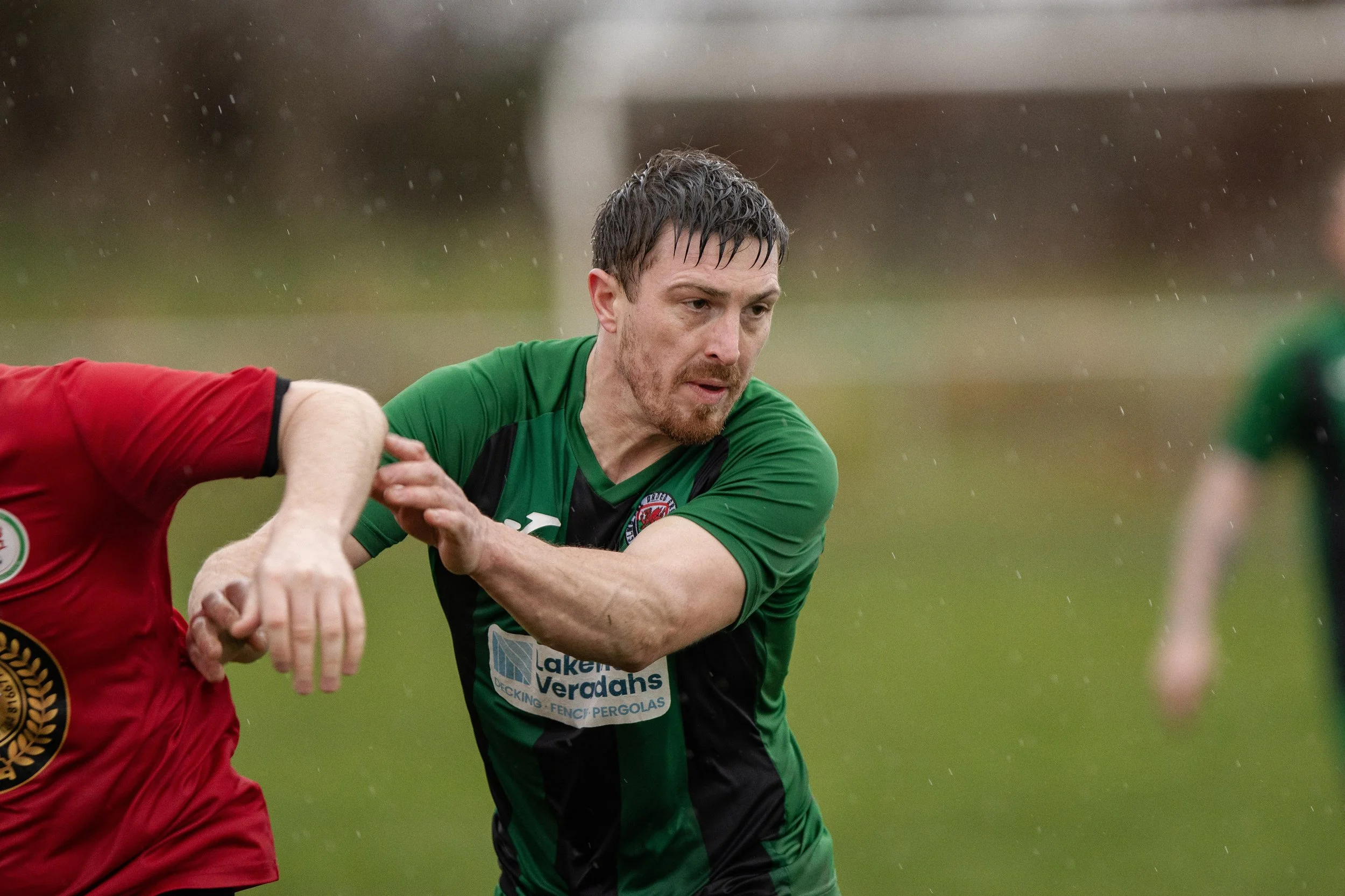A rugby player wearing a green and black jersey on a rainy field, reaching out with his left arm towards a player in a red jersey.
