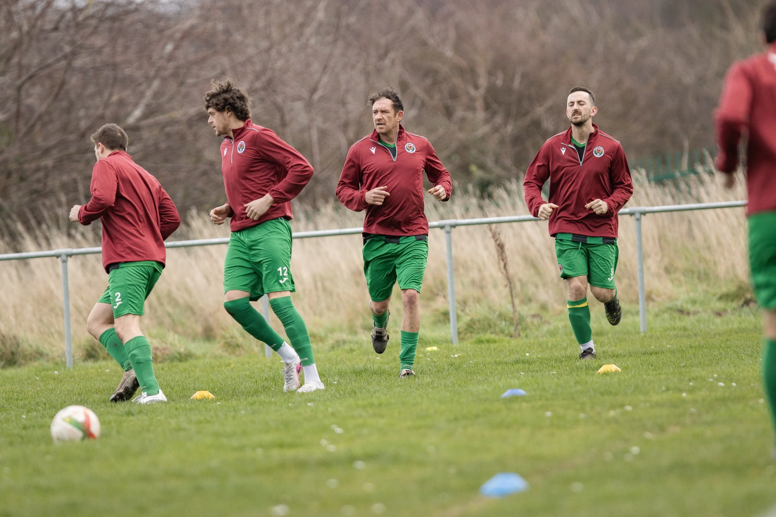 Soccer players in red jackets and green shorts jogging on practice field with cones.