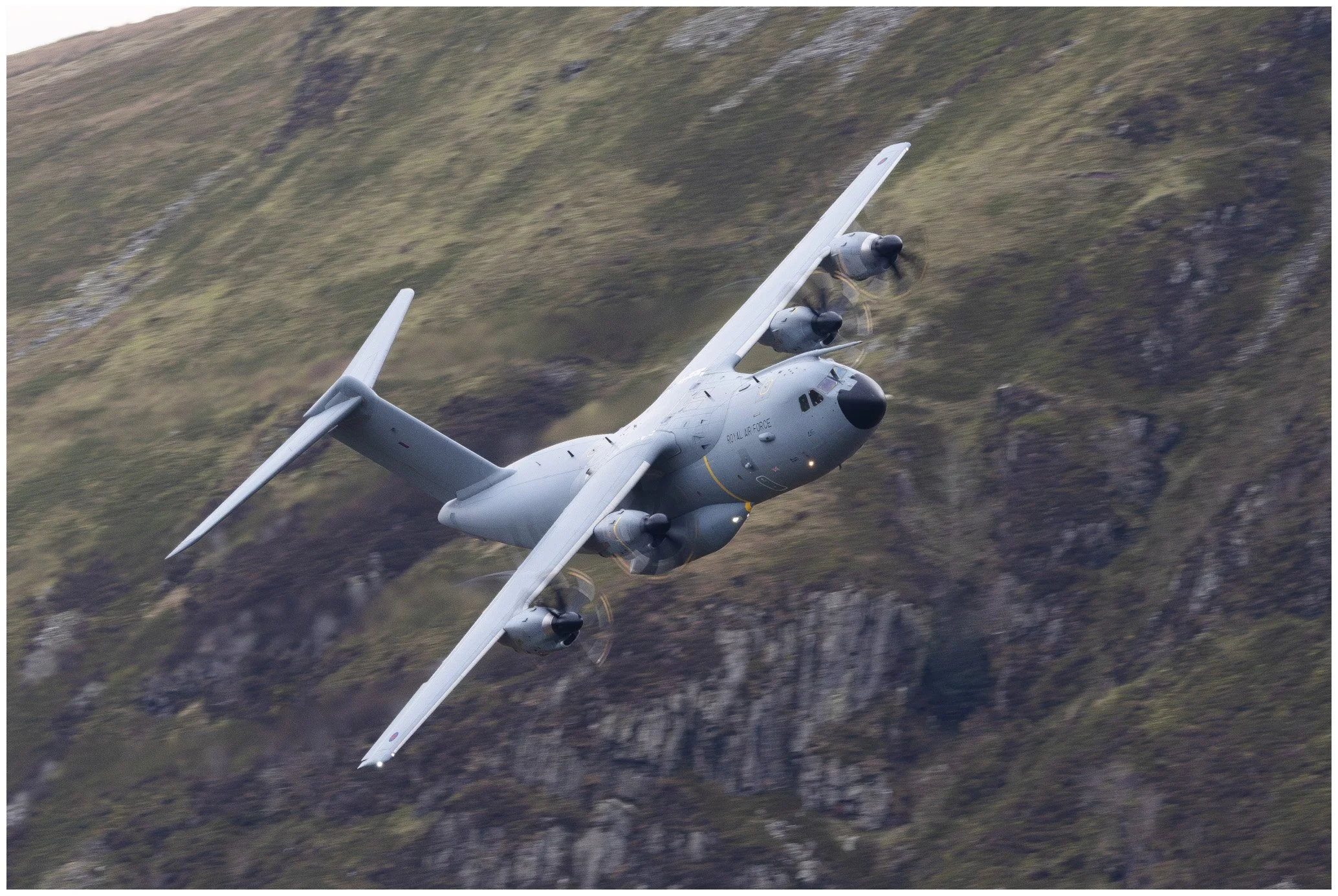 A military transport aircraft flying at an angle over a mountainous landscape.