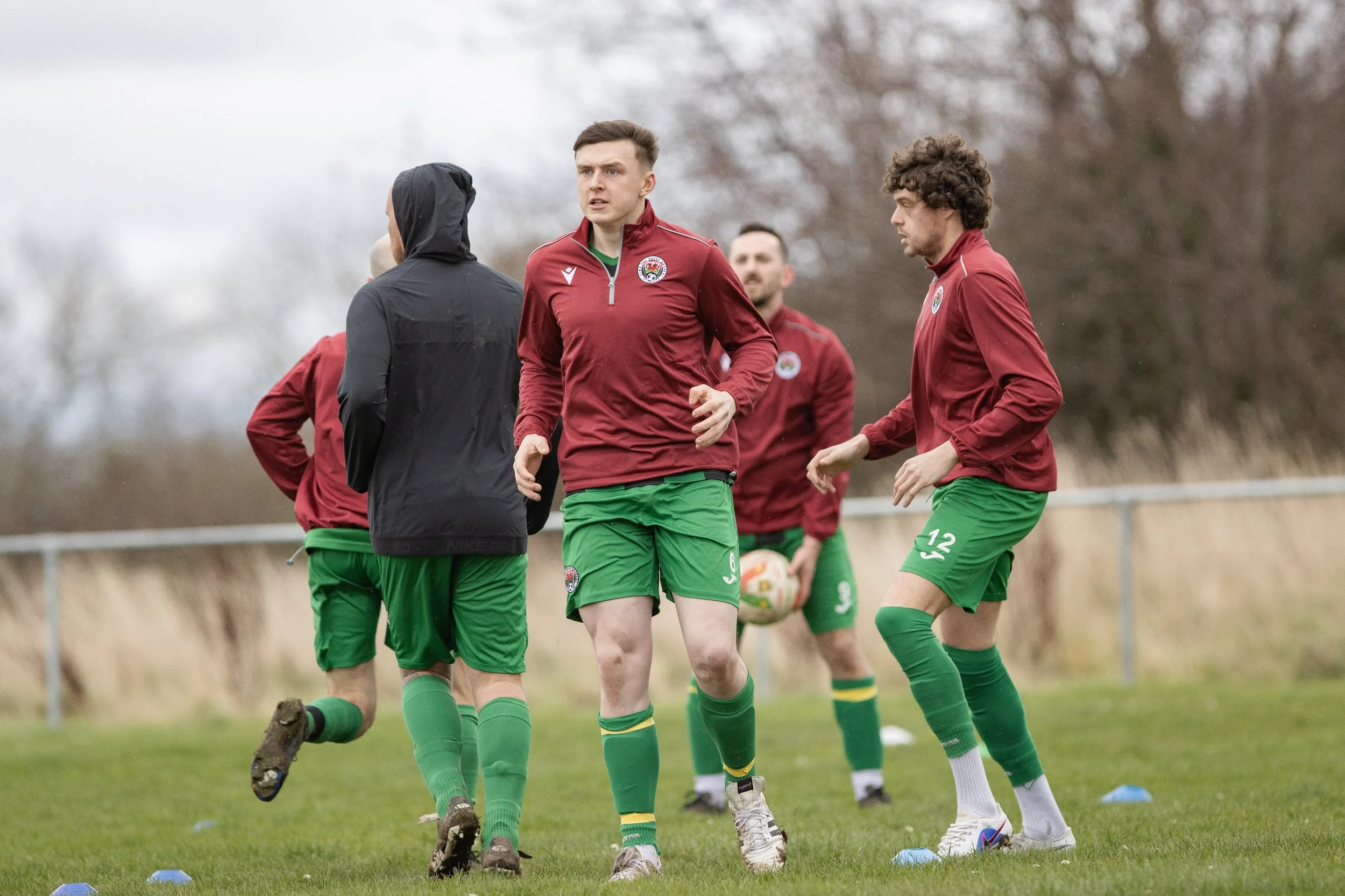Soccer players in red and green uniforms practicing on a grassy field with trees in the background.