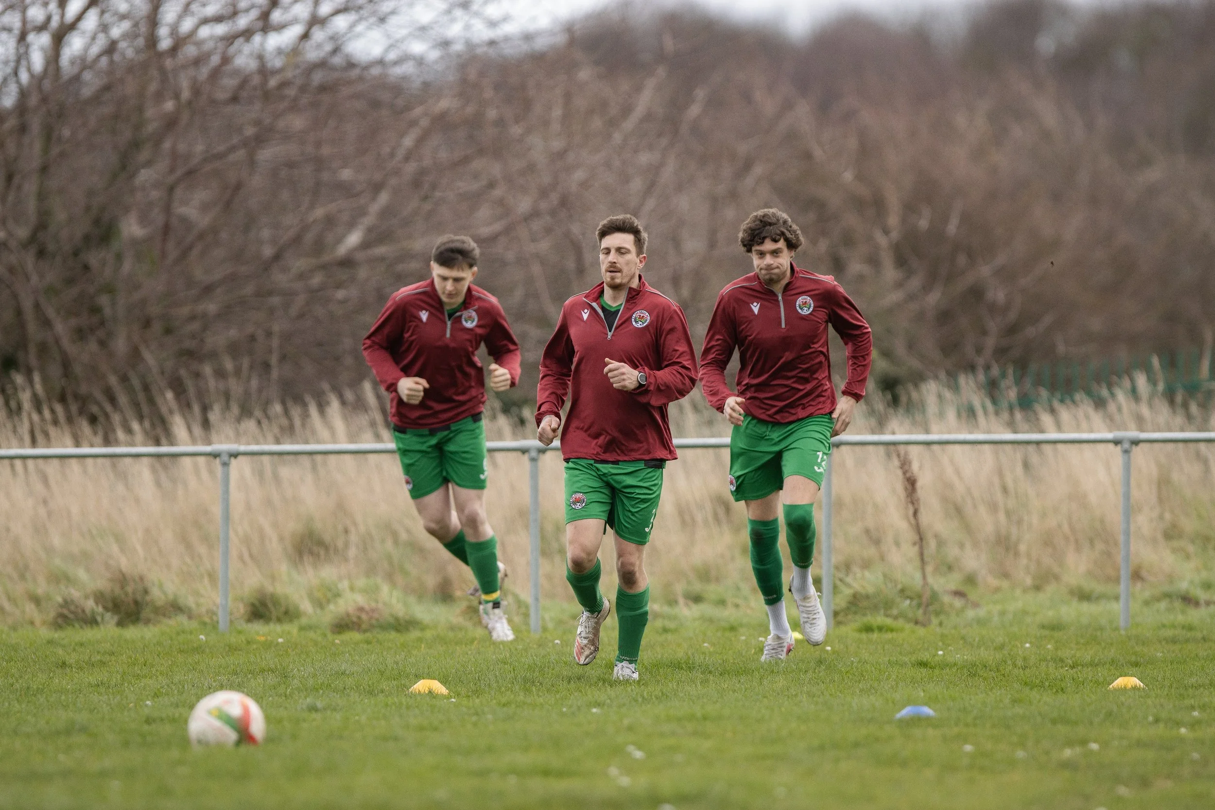 Three soccer players in red jackets and green shorts jogging on a grassy field during training with trees in the background.
