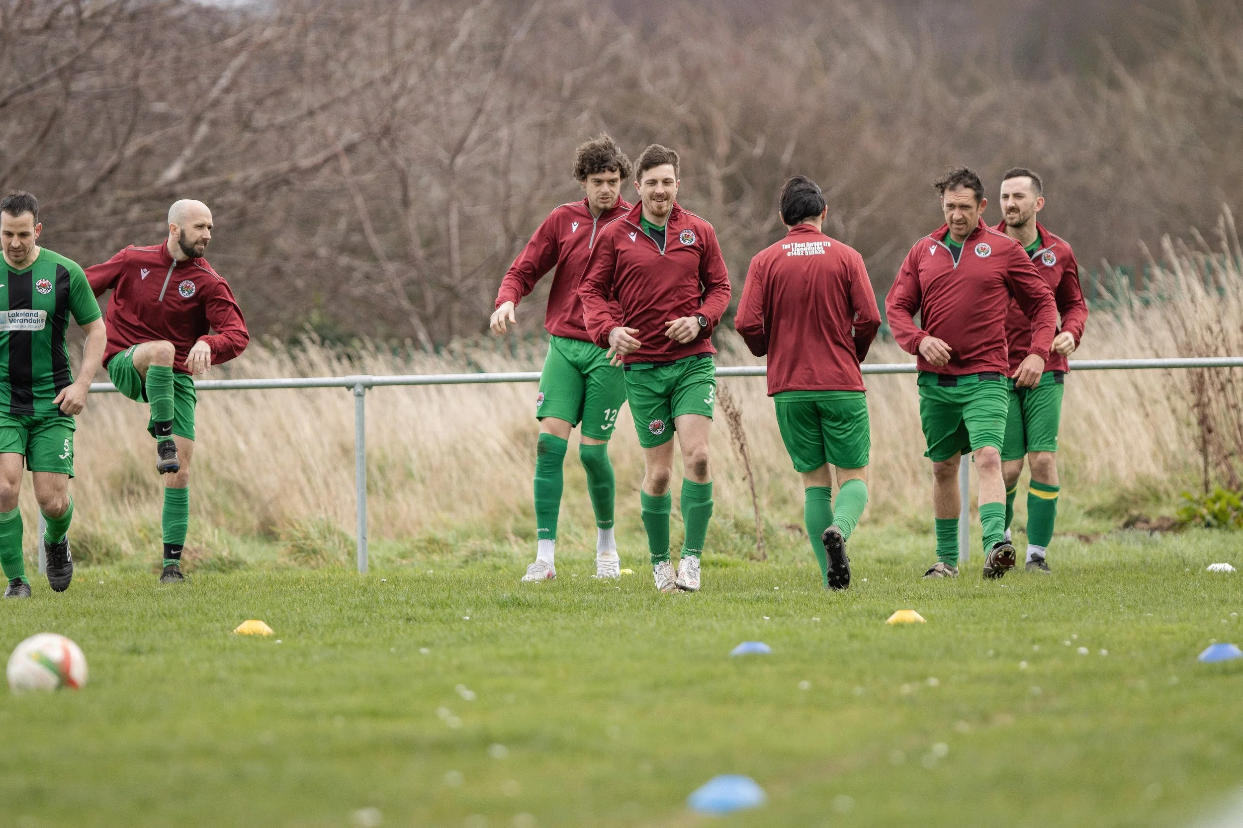 A group of soccer players warming up on a grassy field, wearing red and green uniforms, with some stretches and exercises, surrounded by markers and natural scenery.