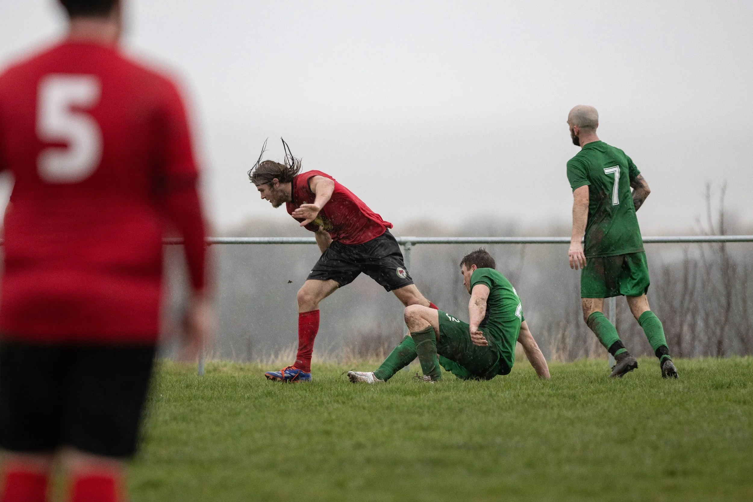 A soccer match in rainy weather, with players in red and green jerseys actively competing on the field. A player in red is falling forward after a tackle from a player in green who is seated on the grass, and two other players in green nearby.