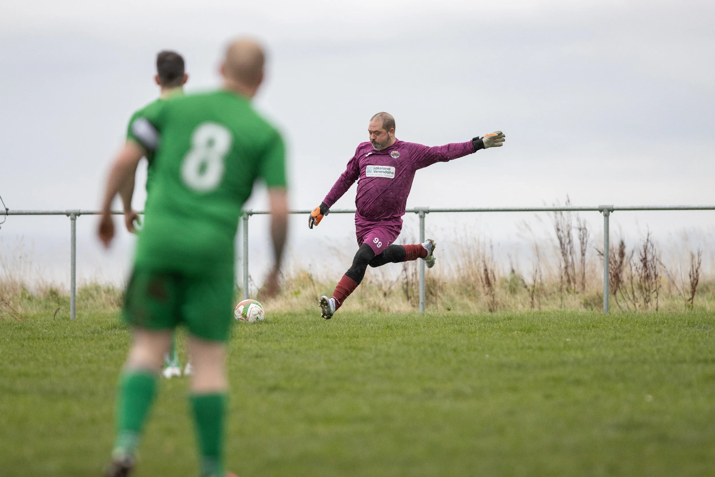 A soccer goalkeeper in a purple uniform kicking a soccer ball on a grassy field during an outdoor game.