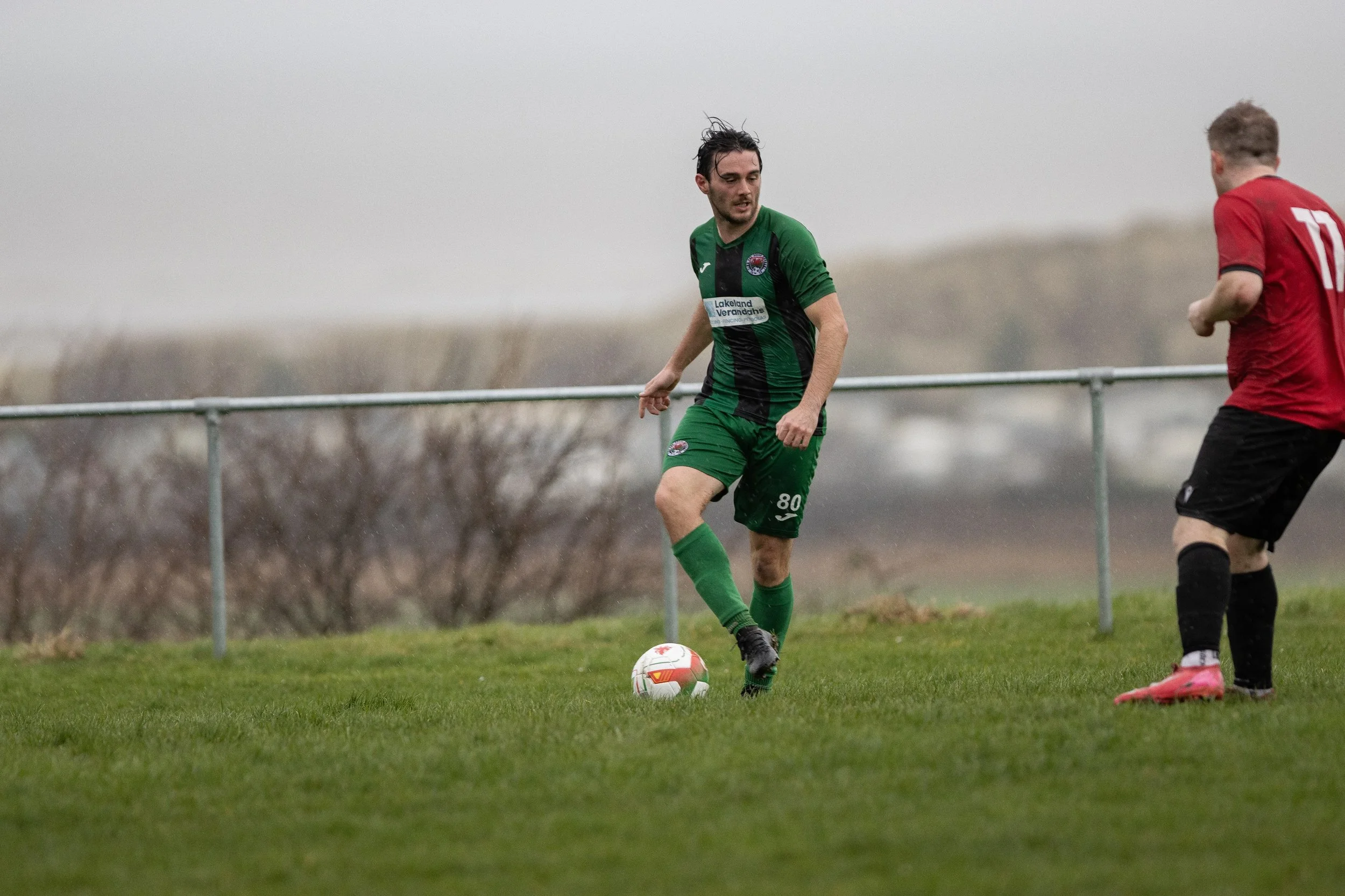 A soccer player in a green and black uniform preparing to kick a soccer ball on a grassy field, with another player in a red and black uniform nearby, under a cloudy sky.