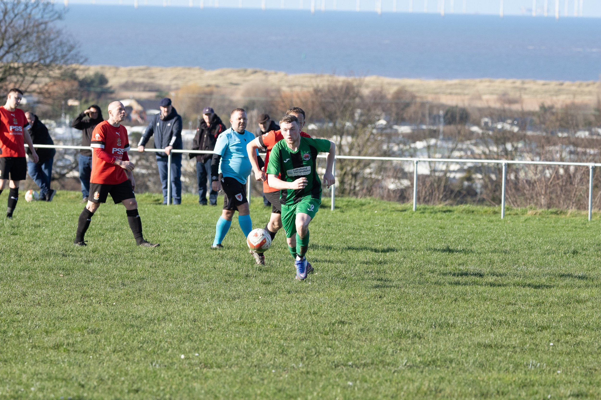 Soccer game on grassy field with players in red, green, and blue uniforms and spectators in the background, overlooking a landscape with trees, buildings, and water.