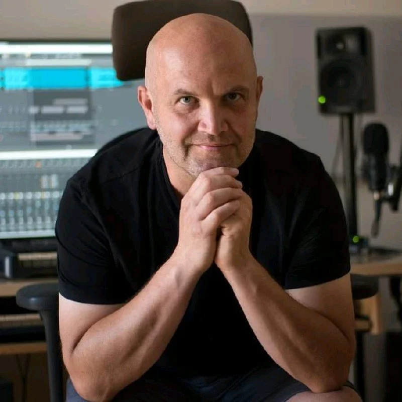 Der bekannte Komponist Jochen Flach. Mann in einem Tonstudio vor einem Computer mit Musiksoftware, sitzt auf einem Stuhl, hat den Kopf leicht gesenkt, hält die Hände vor das Kinn und blickt direkt in die Kamera.