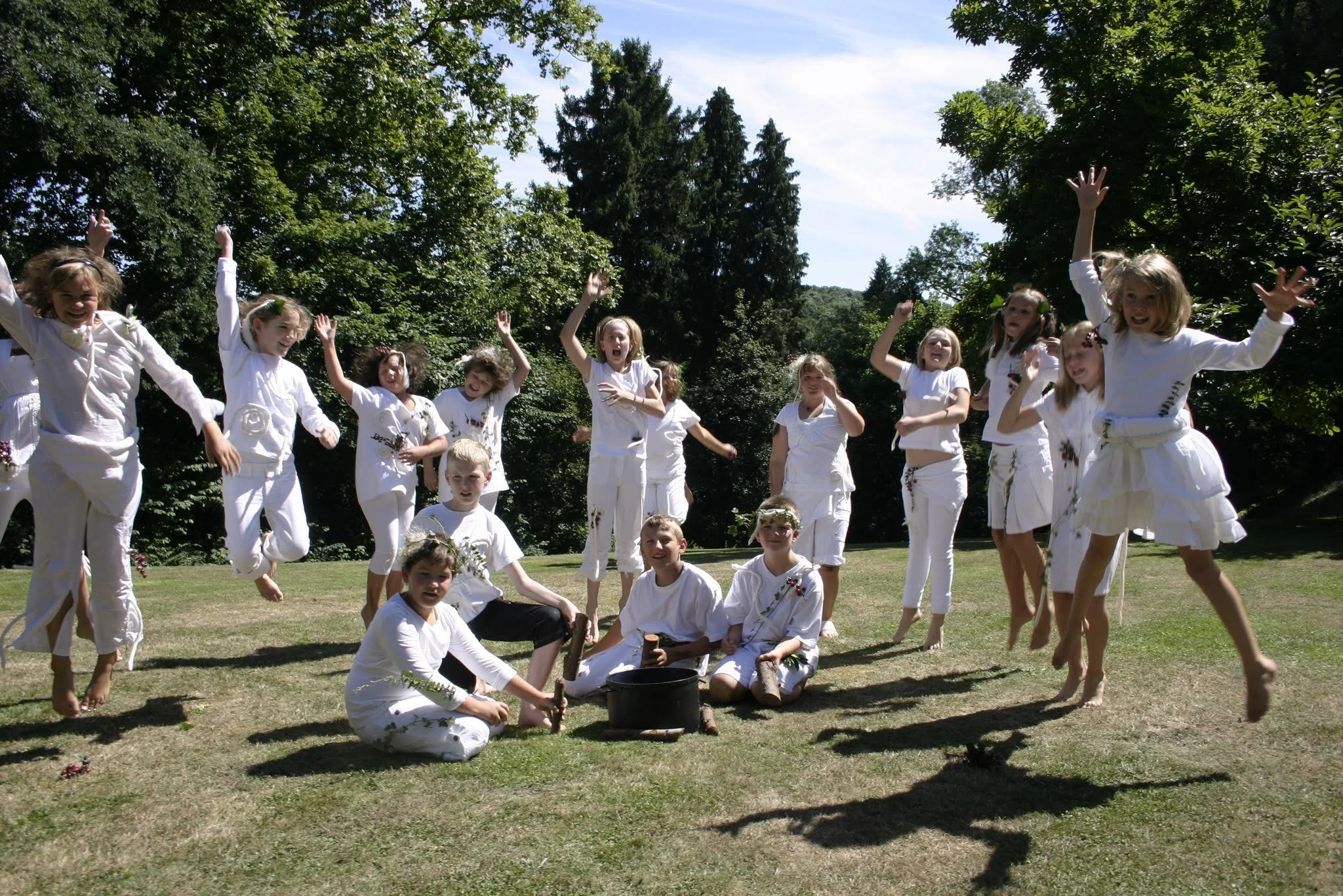 Kinder in weißen Kleidern springen fröhlich auf einer Wiese im Freien, umgeben von Bäumen.