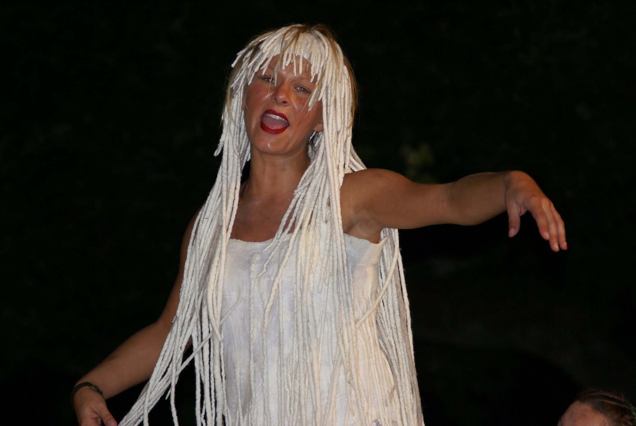 Eine Frau mit weißen Dreadlocks trägt ein weißes Oberteil und scheint beim Singen oder Sprechen zu sein.