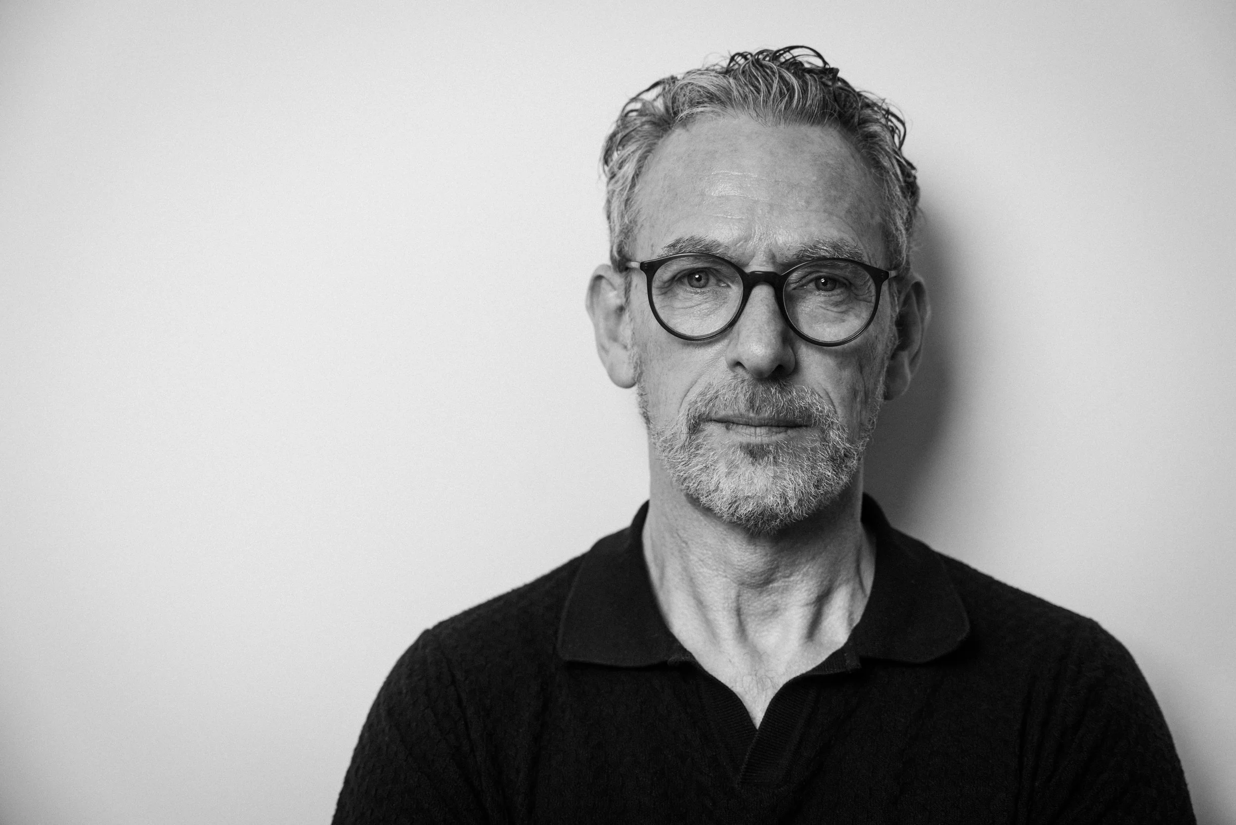 Schwarz-Weiß-Fotografie eines älteren Mannes mit Locks und Brille, trägt einen schwarzen Pullover, steht vor weißer Wand.