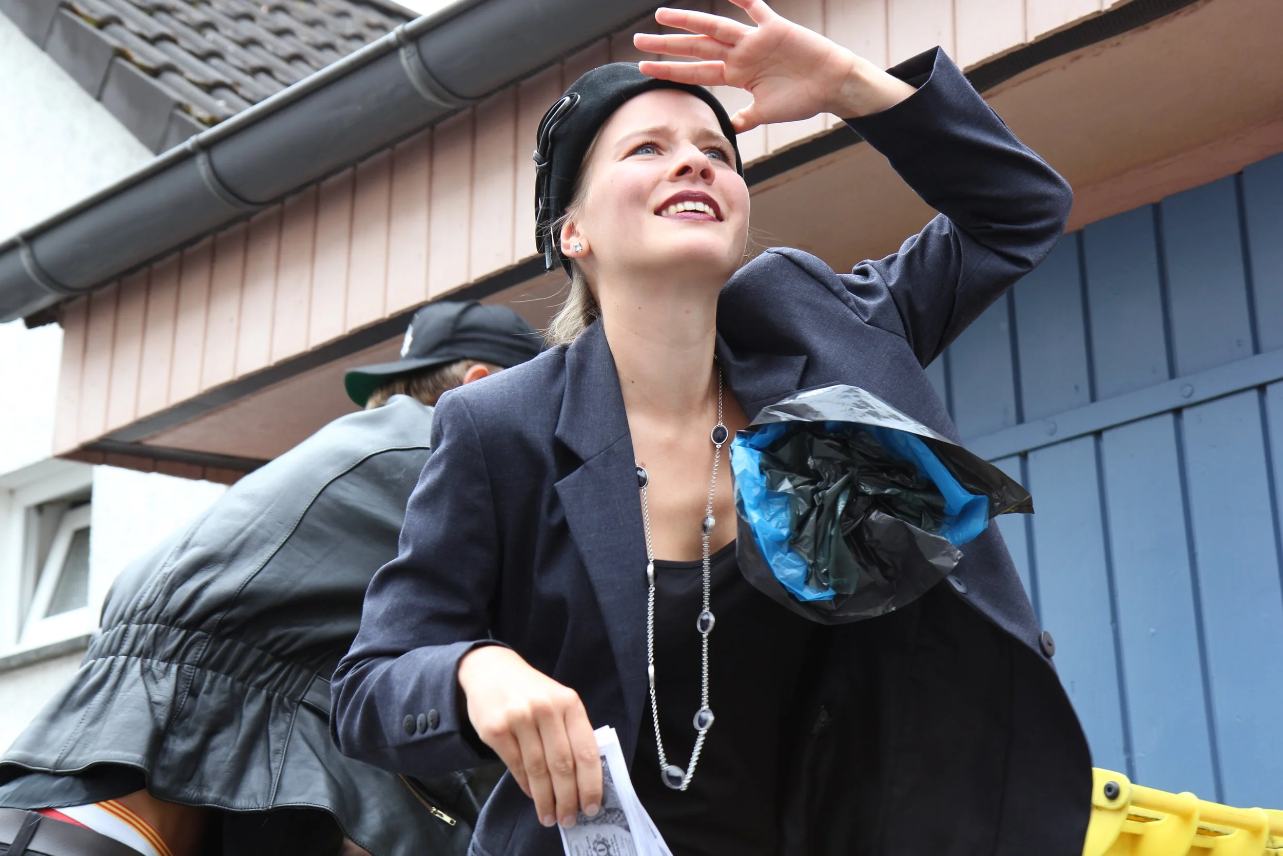 Junge Frau mit dunkler Jacke und schwarzem Hut hält ihre Hand über die Augen und schaut nach oben, während sie neben einem Mann steht, der einen schwarzen Lederjacke trägt. Sie steht vor einem Gebäude mit blauer und brauner Fassade.