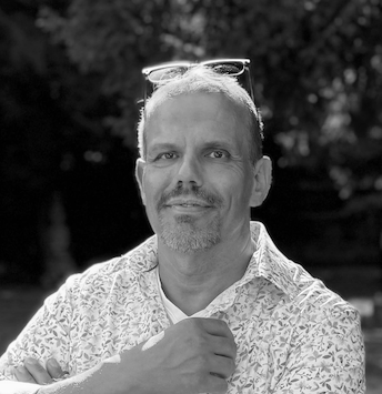 Ein Mann mit kurzen Haaren und einem Bart, trägt eine Sonnenbrille auf dem Kopf und ein Hemd, steht im Freien mit Bäumen im Hintergrund.