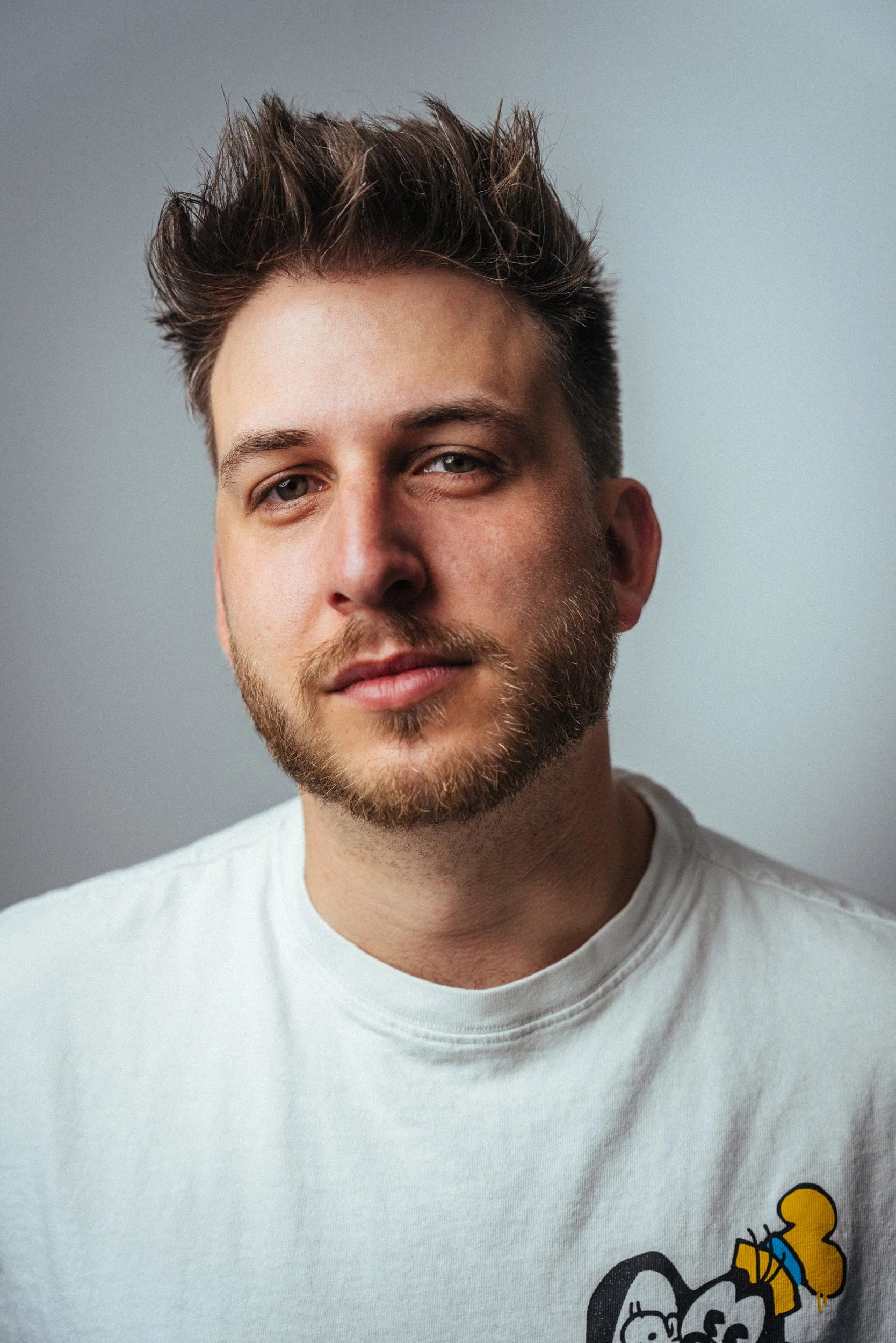 Nahaufnahme eines jungen Mannes mit braunem, gewelltem Haar und Bart, trägt ein weißes T-Shirt mit einem gelben und schwarzen Comic-Design. Leon Roskoni  in  Das Wilde Weib – Das Musical · Birsteiner Festspiele 2027