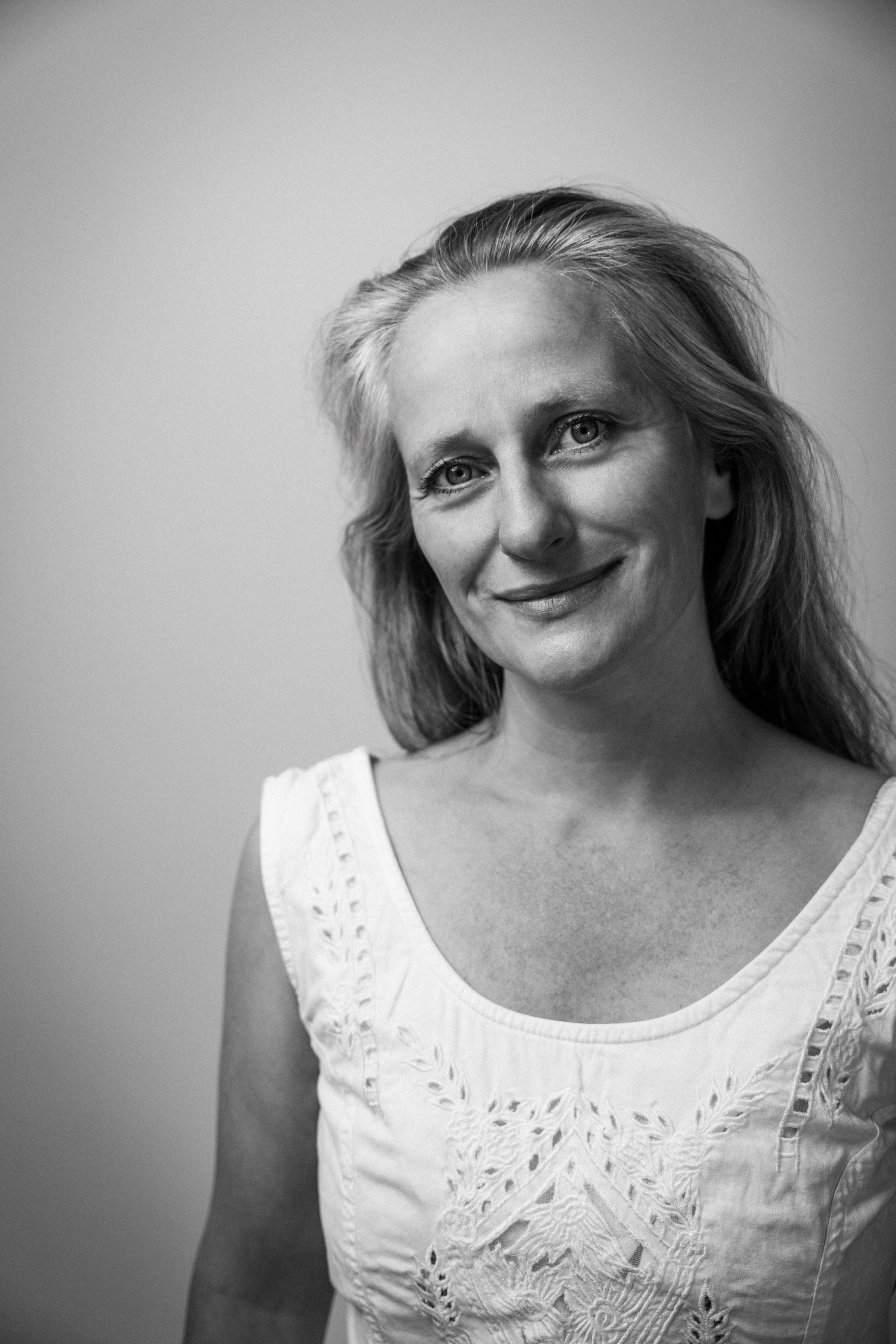 Schwarz-Weiß-Fotografie einer lächelnden Frau mit mittelblondem Haar, die ein weißes Top mit Stickereien trägt, vor einem neutralen Hintergrund.
