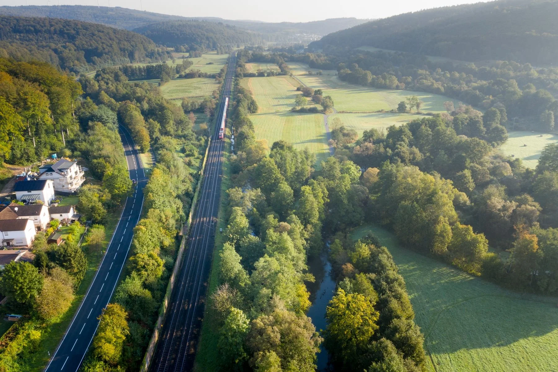 Luftaufnahme eines grünen, ländlichen Gebiets mit Wäldern, Feldern, einem Fluss und einer Bahnlinie, die durch die Landschaft führt.