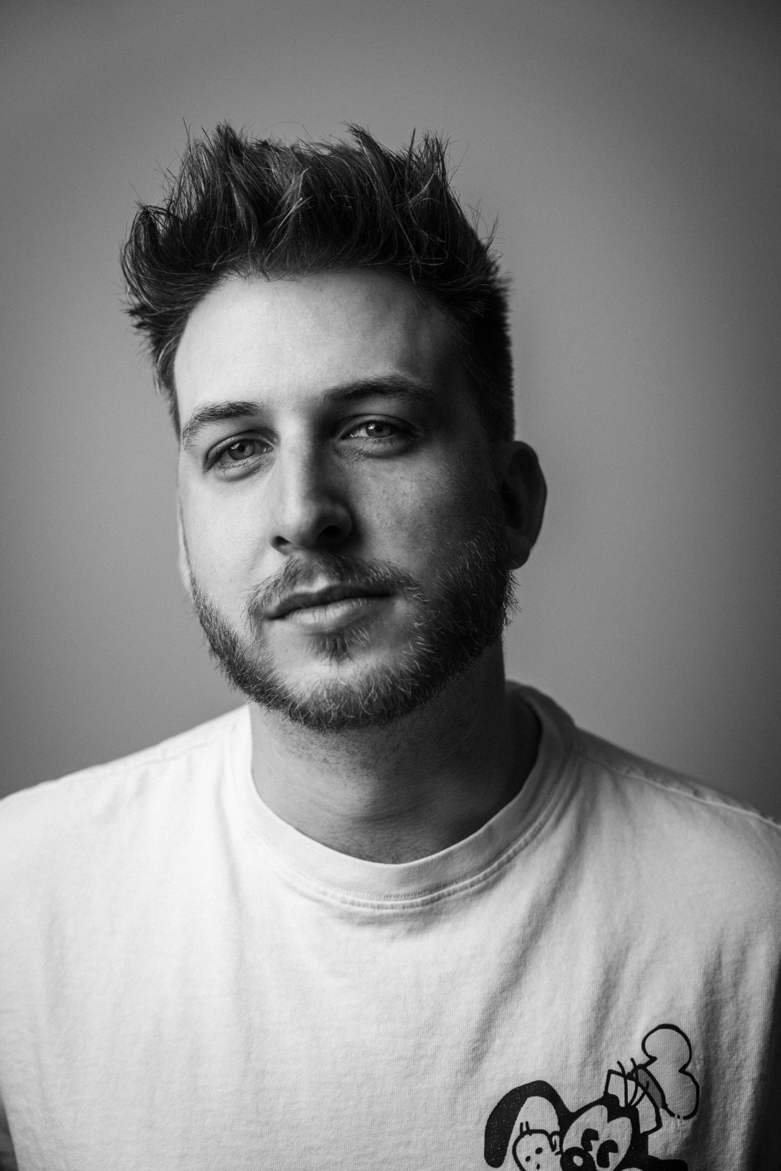 Schwarz-weiß-Fotografie eines jungen Mannes mit charmantem Gesichtsausdruck, kurze Haare und Vollbart. Leon Roskoni Intendant.  in  Das Wilde Weib – Das Musical · Birsteiner Festspiele 2027