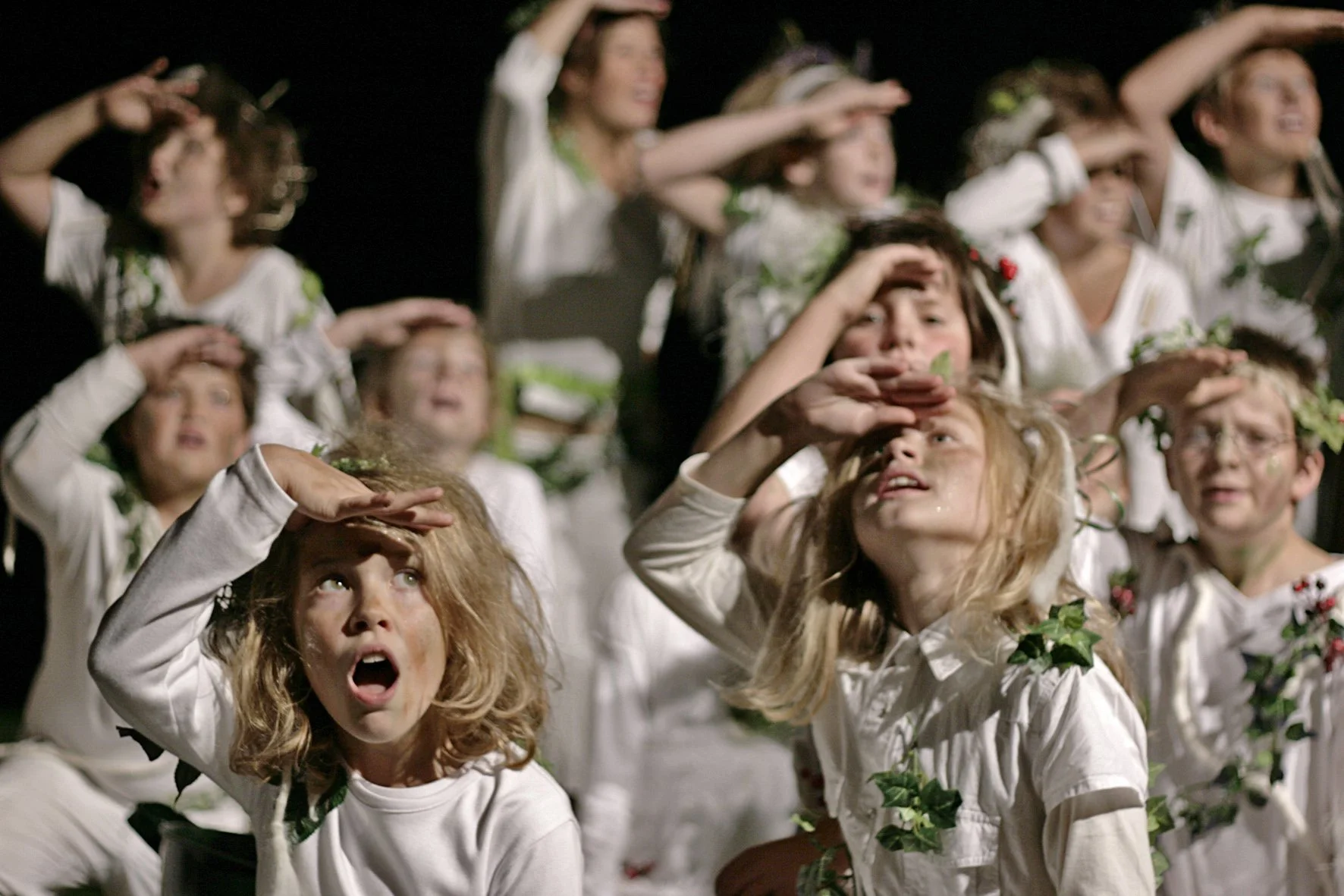 Kinder in weißen Kostümen mit grünen Umweltsymbolen, die die Erde repräsentieren, halten die Hand an die Stirn und schauen in die Ferne bei einer Inszenierung oder Aufführung.