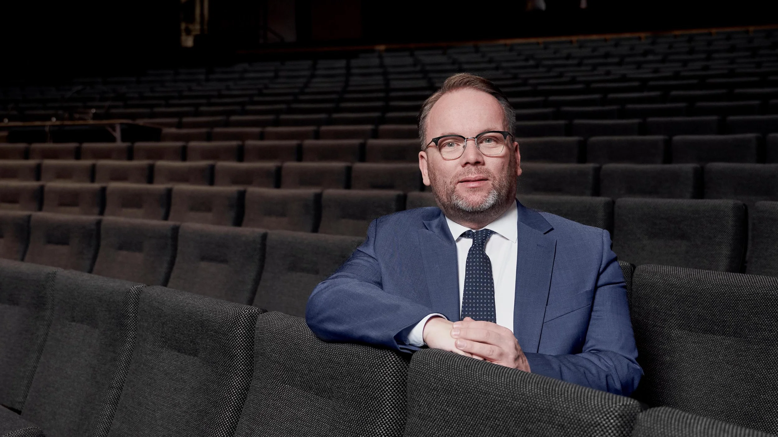 Ein Mann in einem blauen Anzug und mit Brille sitzt in einem leeren Theater oder Auditorium, die Sitze sind grau. Er schaut nach vorne, die Armen sind verschränkt.