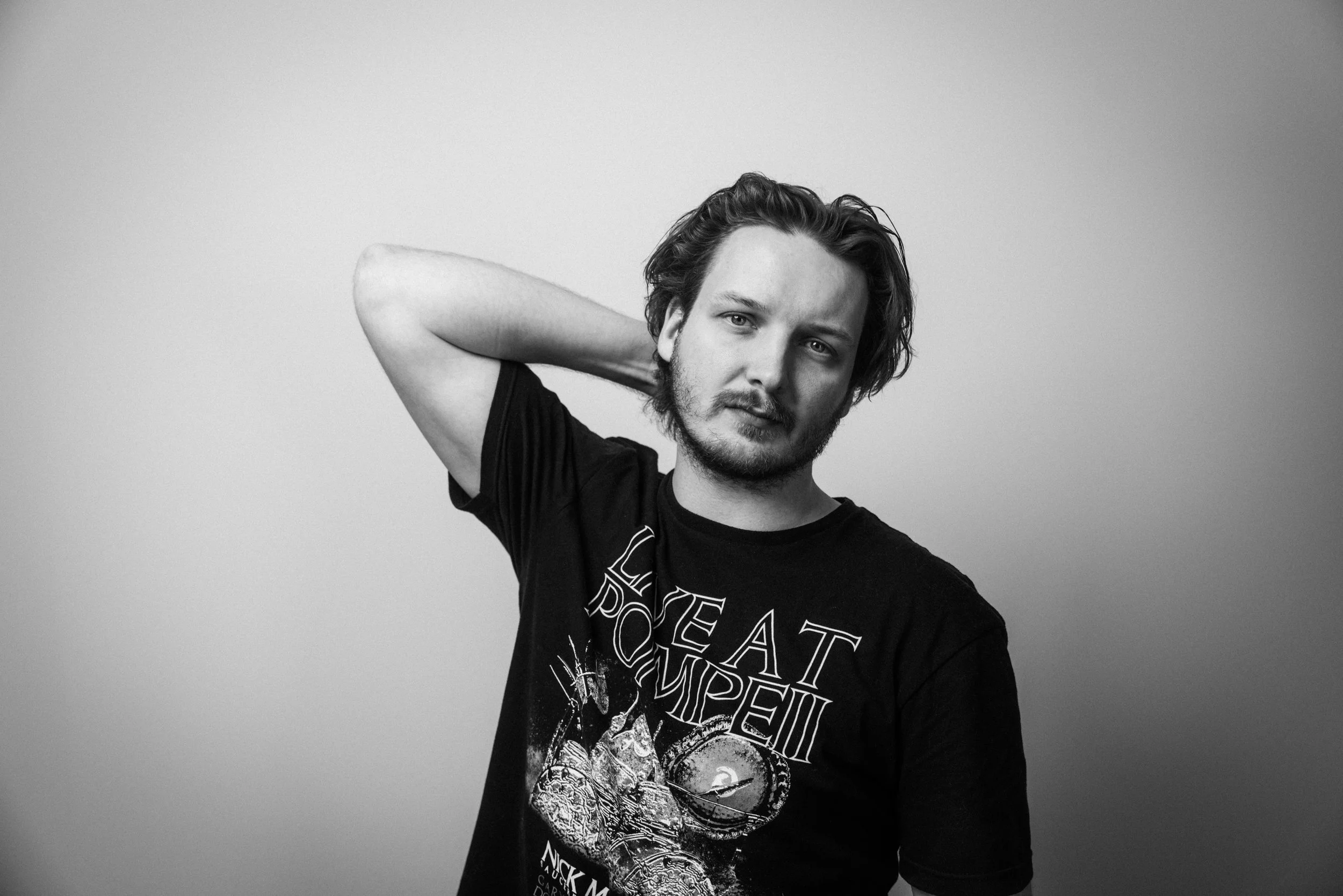 Schwarziges Portrait eines jungen Mannes mit langen, kletten Haaren, der eine Hand hinter den Kopf legt und auf die Kamera schaut, gegen einen einfachen hellgrauen Hintergrund.