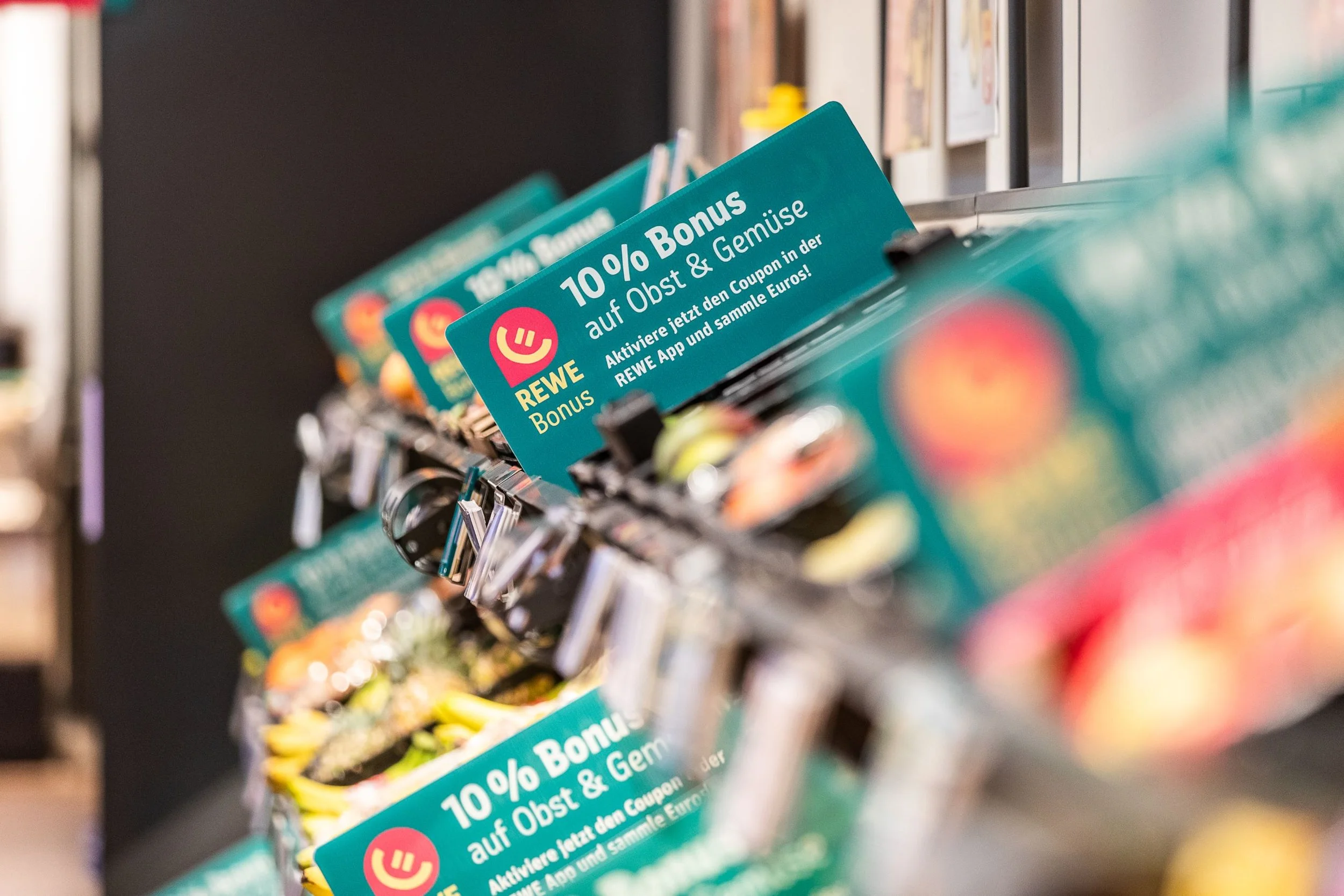 Werbeplakat in einem Supermarkt für einen Rabatt von 10% auf Obst und Gemüse bei REWE, mit Aufforderung, die REWE App zu aktivieren, um den Coupon zu sammeln.