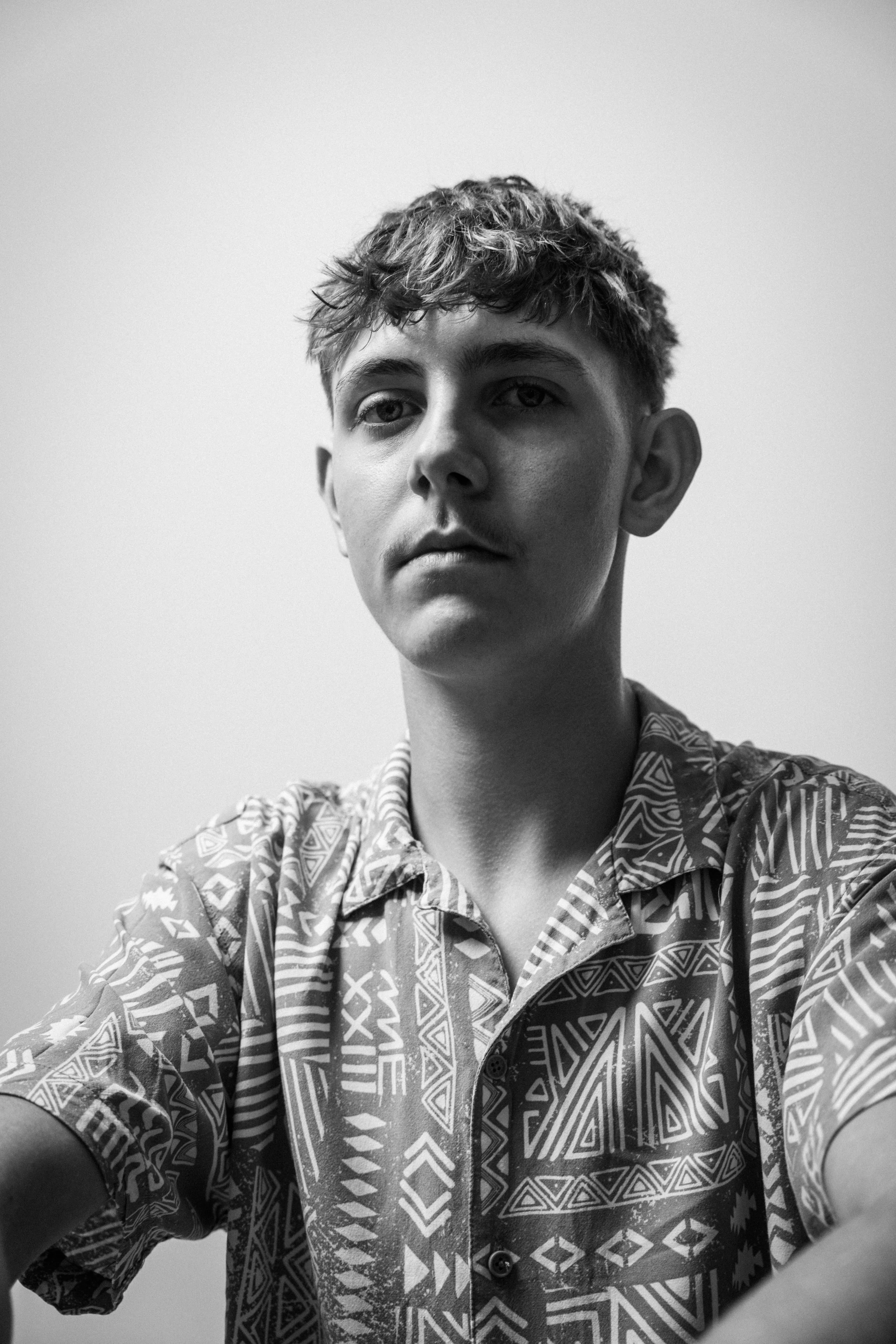 Schwarz-Weiß-Foto eines jungen Mannes mit kurzen, lockigen Haaren, der eine bunte, geometrisch bedruckte Hemd trägt, blickt ernst in die Kamera.