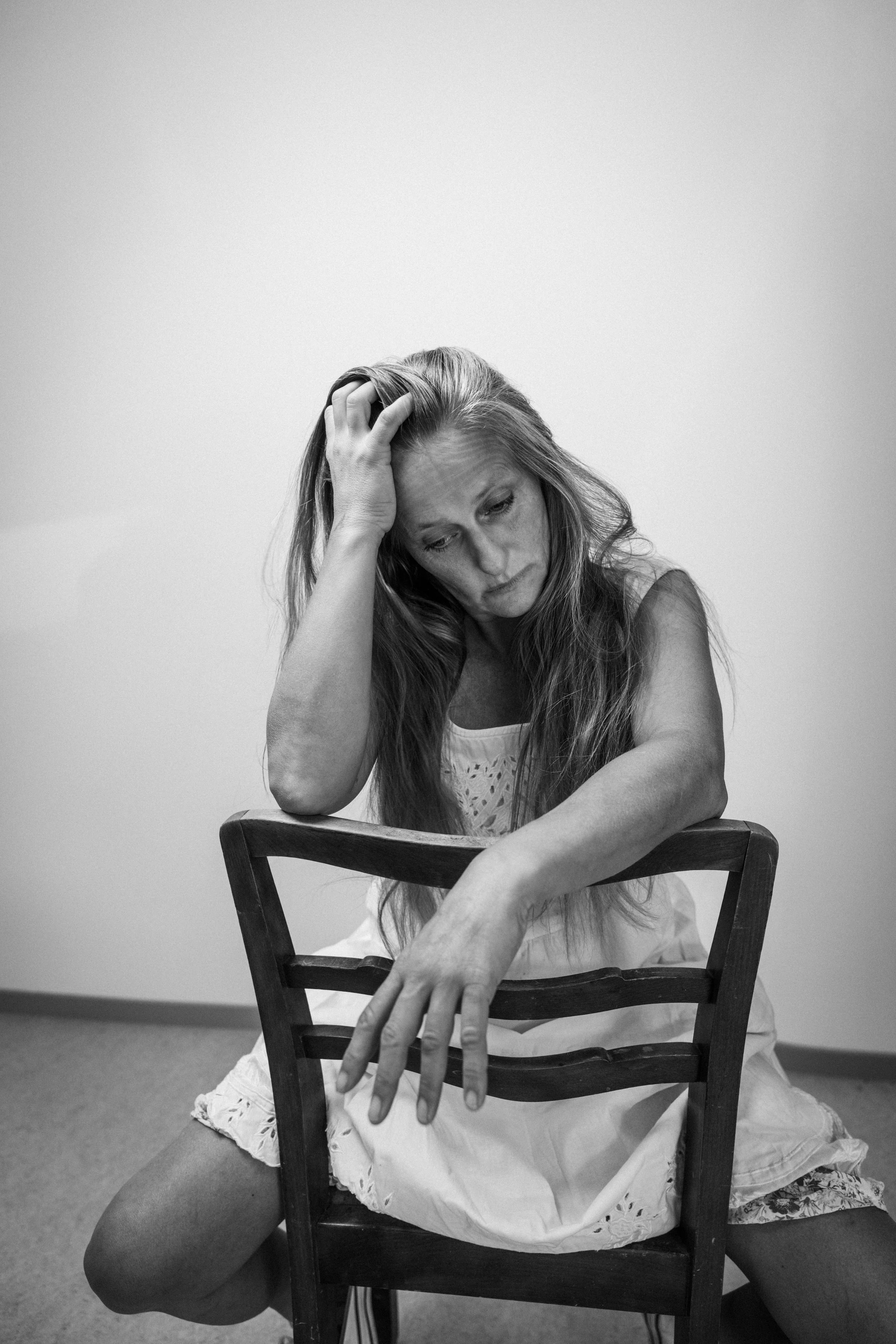 Eine Frau sitzt auf dem Boden mit einem hölzernen Stuhl um sie herum, sie sieht nach unten und hat eine Hand am Kopf, während die andere nach vorne reicht. Das Bild ist in Schwarz-Weiß gehalten.