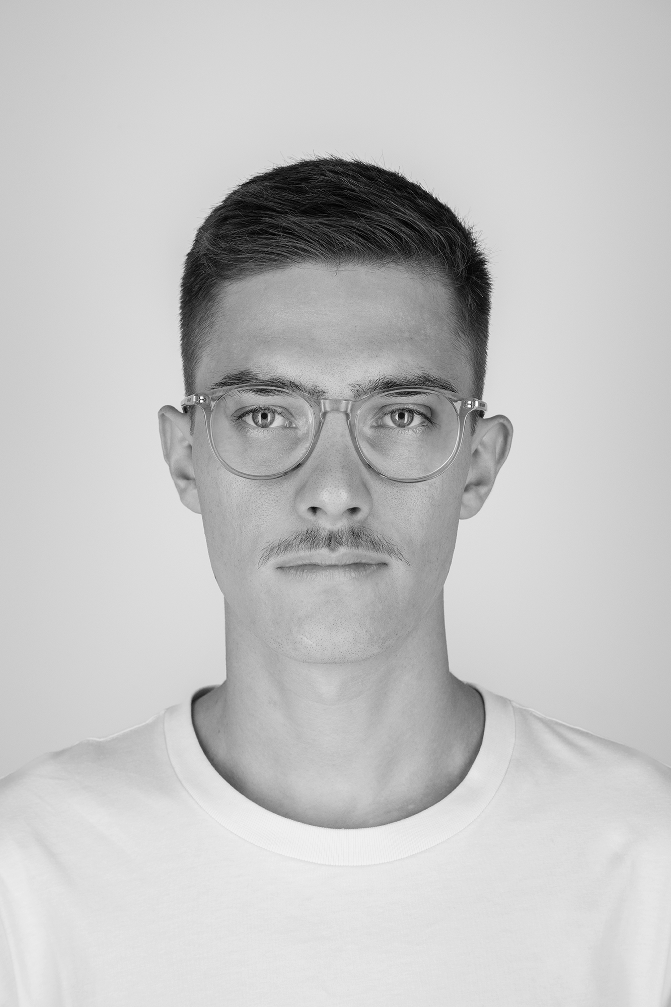 Schwarz-weiß Foto eines jungen Mannes mit kurzen dunklen Haaren, Brille und weißem T-Shirt vor hellem Hintergrund.