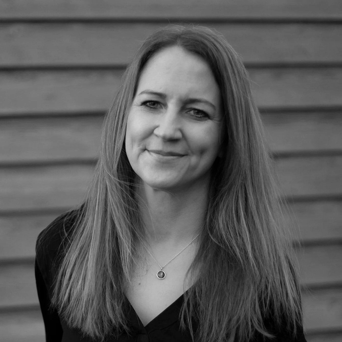 Schwarz-weiß-Fotografie einer Frau mit langen, glatten Haaren vor einer Holz-Wand. Sie trägt eine Halskette und lächelt leicht.