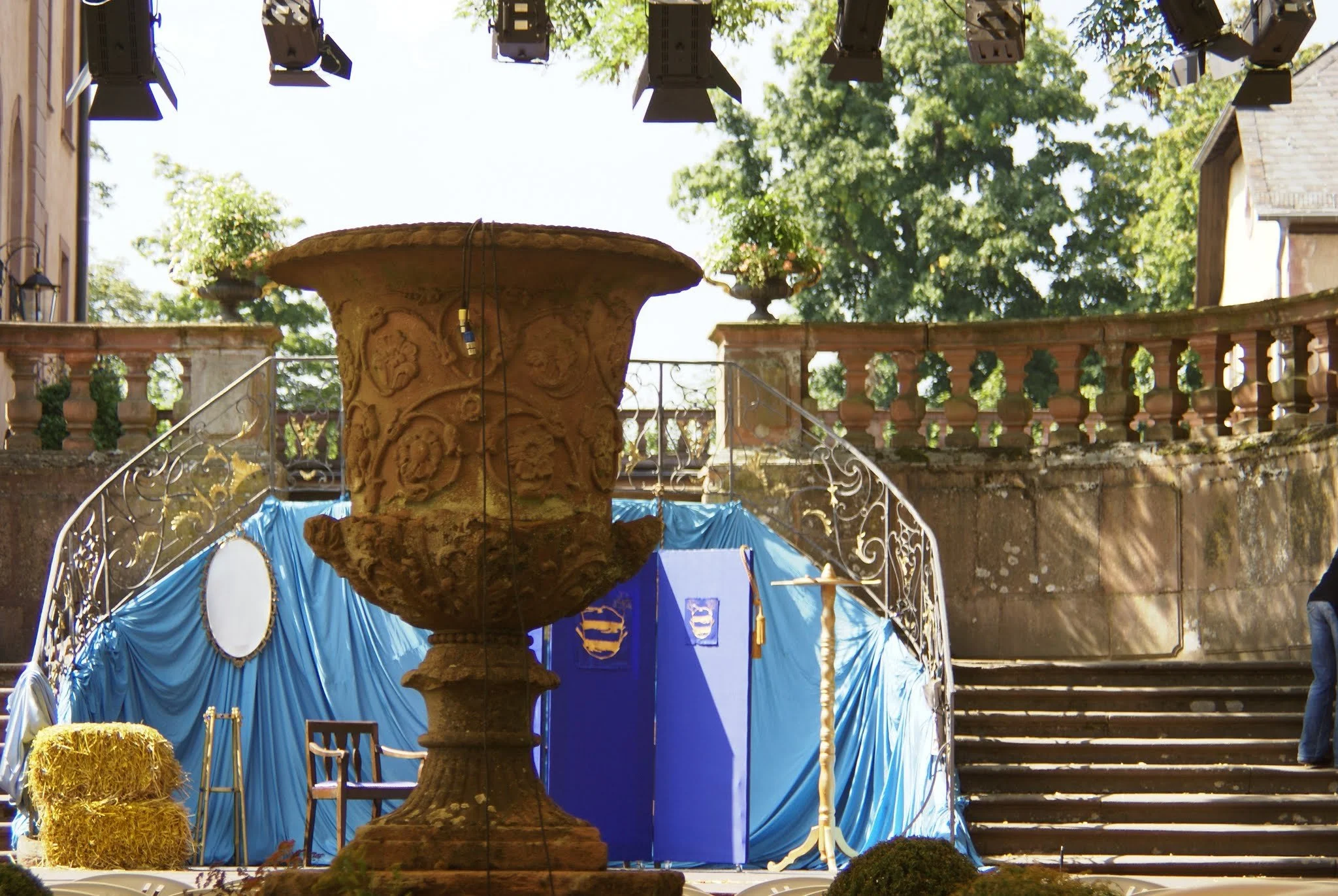 Bühne mit blauer Dekoration, alte steinerne Brunnen im Vordergrund, Bühnenlichter hängen über der Bühne, im Hintergrund Bäume und ein alter Balkon mit Säulen.