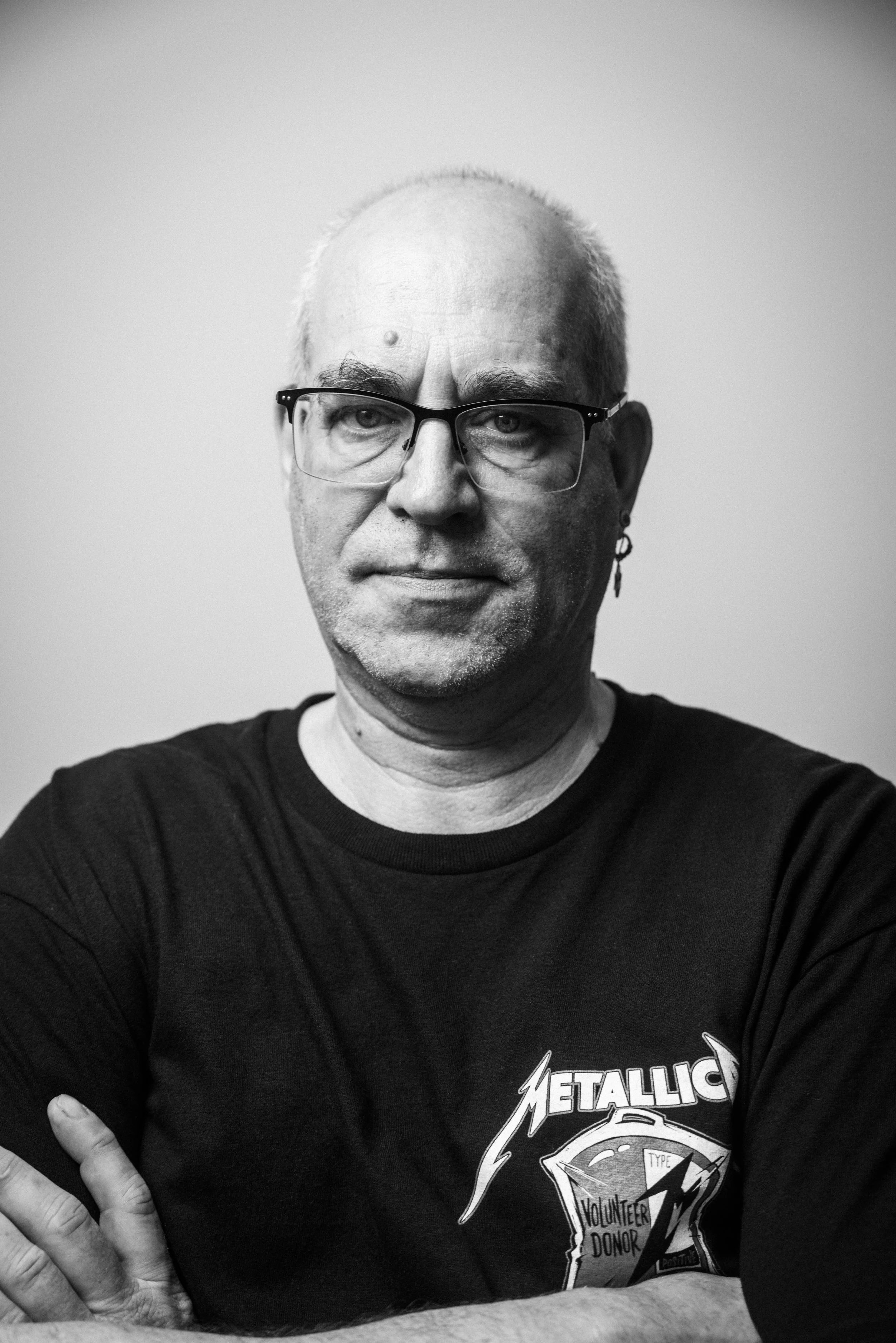 Schwarz-weiß-Fotografie eines Mannes mit Brille, Earring und schwarzen T-Shirt, der die Arme verschränkt, vor einem neutralen Hintergrund.