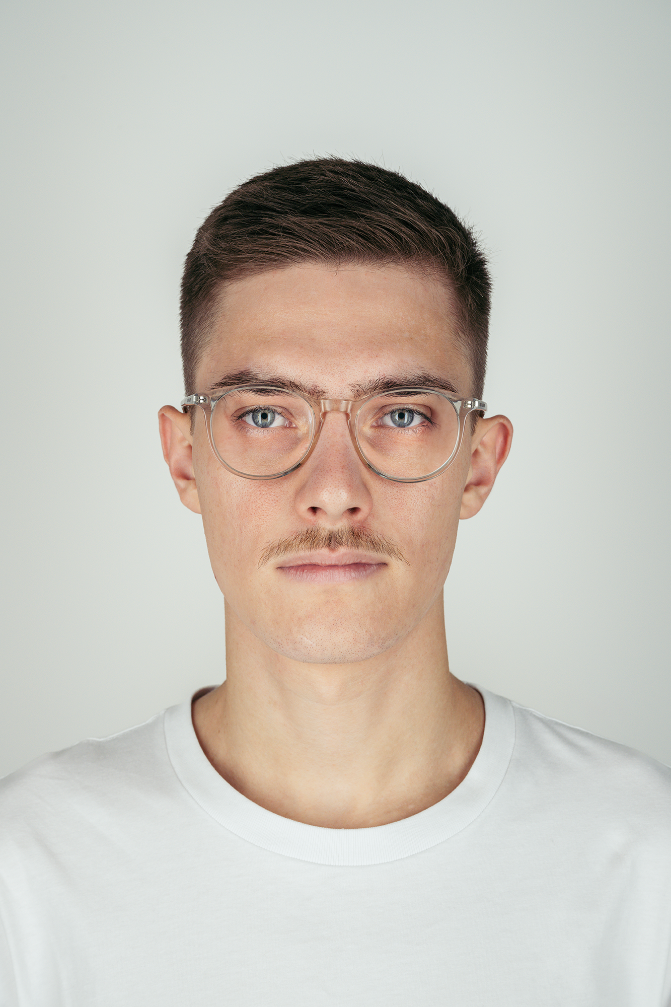 Junger Mann mit kurzen dunklen Haaren, Brille mit transparentem Rand, leichtem Bart und weißen T-Shirt vor einem hellen Hintergrund.