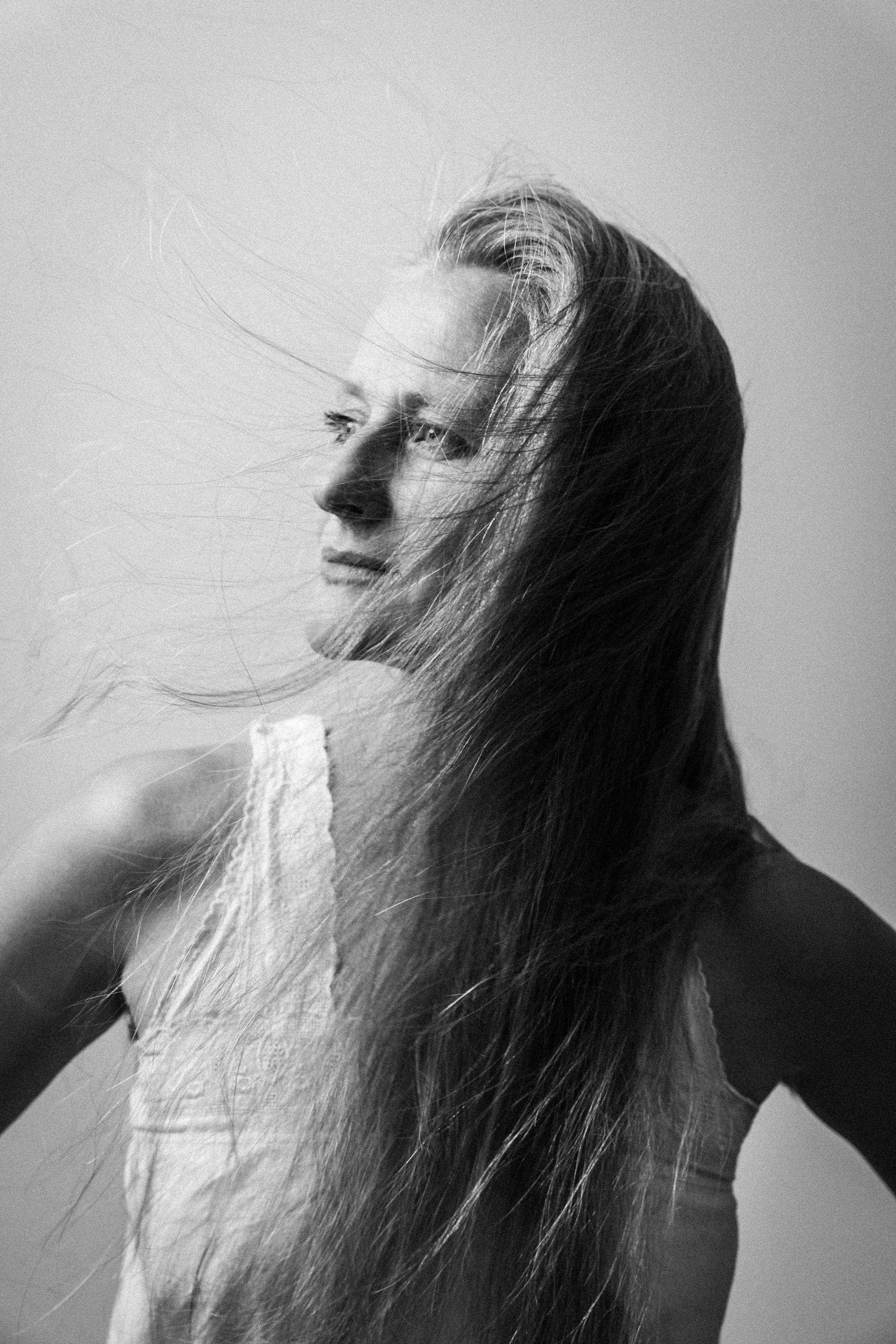 Schwarz-weiß-Fotografie eines jungen Mädchens mit langen Haaren, die vom Wind zerzaust werden, blickt nach links, auf einem neutralen Hintergrund.