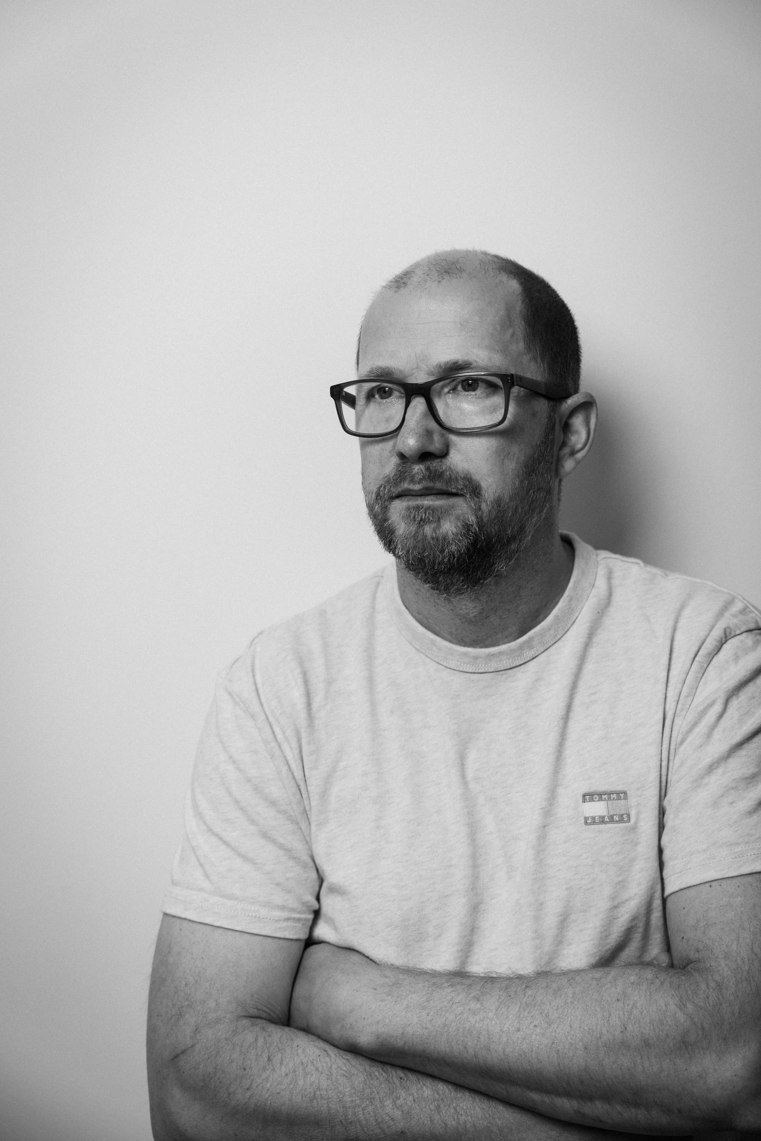Schwarz-weißer Porträt eines Mannes mit Brille, kurzen Haaren und Bart, trägt ein hellgraues T-Shirt mit Tommy Jeans Logo, steht vor einer weißen Wand, Arme verschränkt.