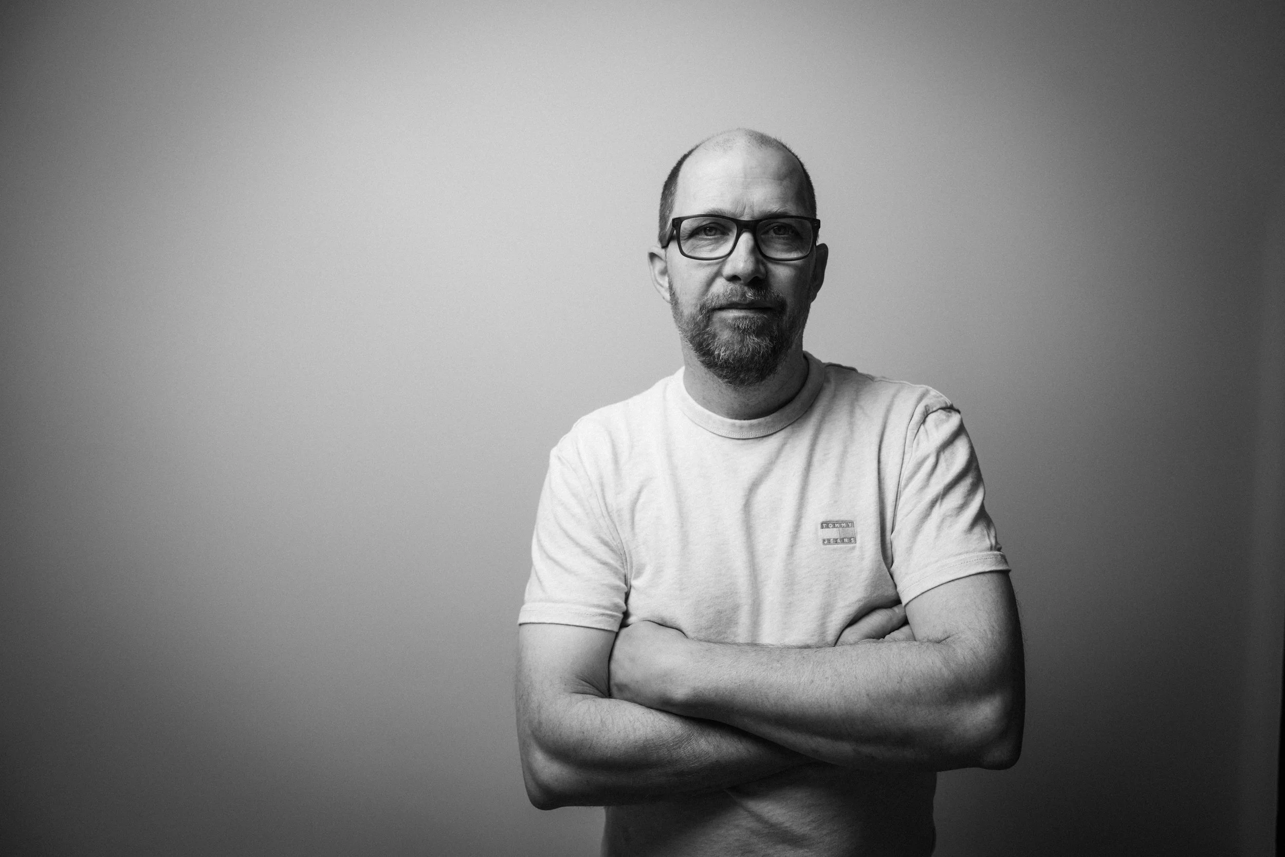 Schwarz-weiß-Fotografie eines Mannes mit Brille, Bart und kurzen Haaren, der ein T-Shirt trägt. Er steht vor einem einfarbigen, hellgrauen Hintergrund und blickt ernst in die Kamera, die Arme verschränkt.