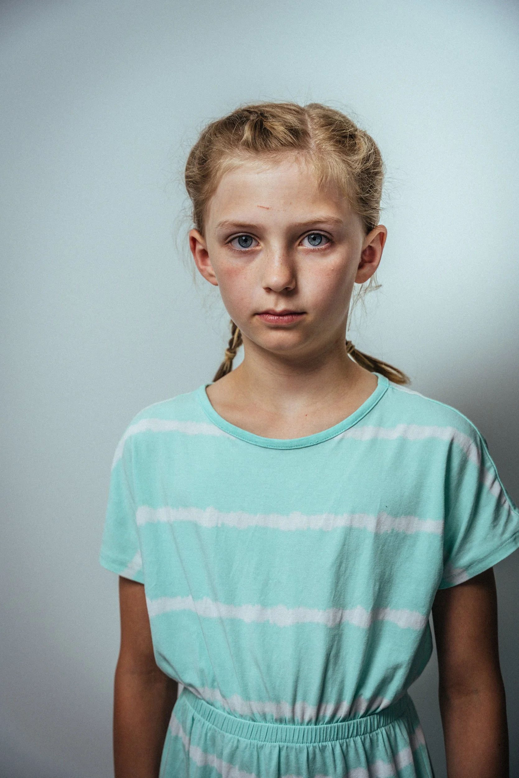 Ein Mädchen mit roten Haaren, blauen Augen und einem Streifen-Shirt in Mint und Weiß, blickt direkt in die Kamera, vor einem grauen Hintergrund.