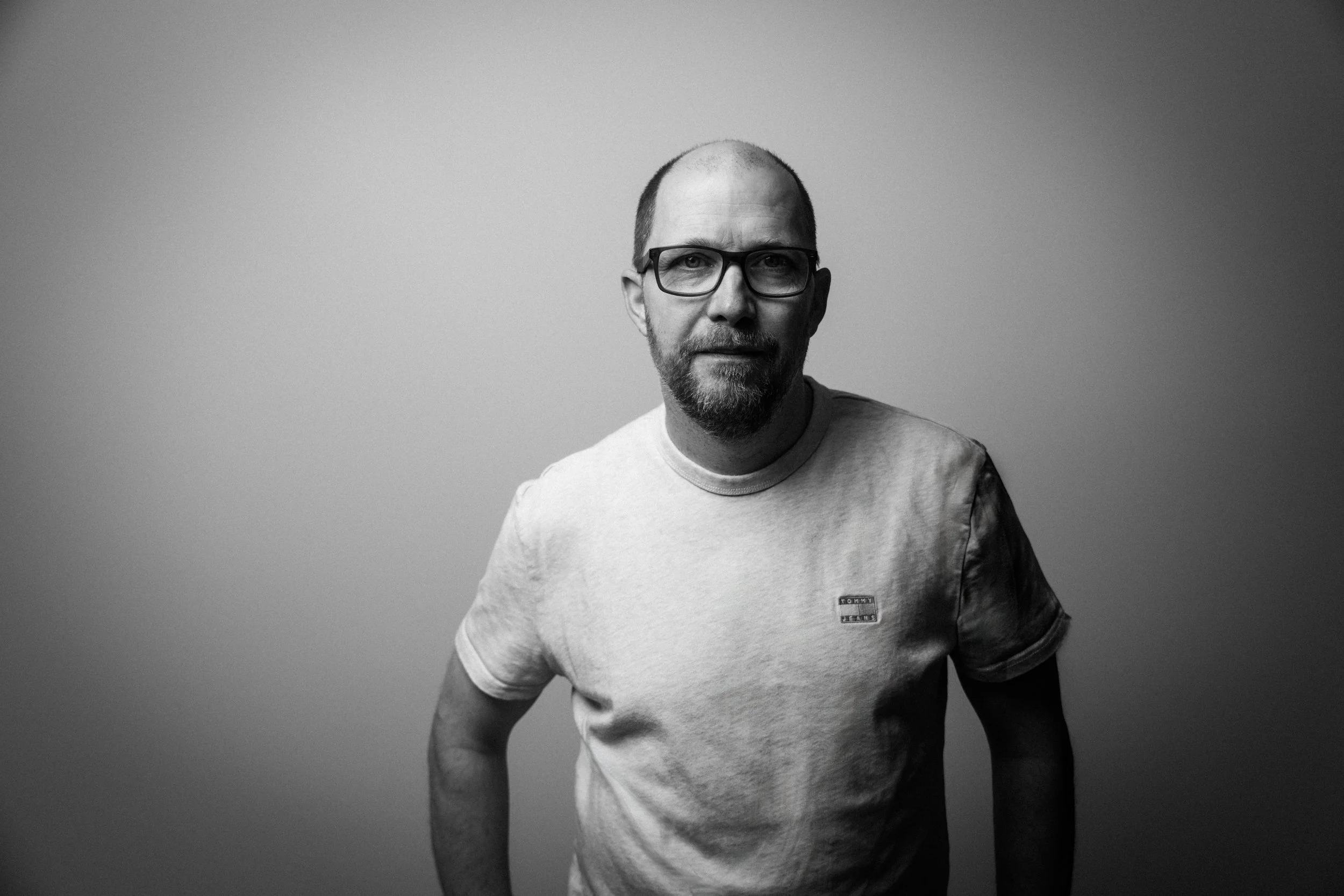 Schwarz-weiße Porträtaufnahme eines Mannes mit Brille, Bart und kurzen Haaren, der ein graues T-Shirt trägt, vor grauem Hintergrund.