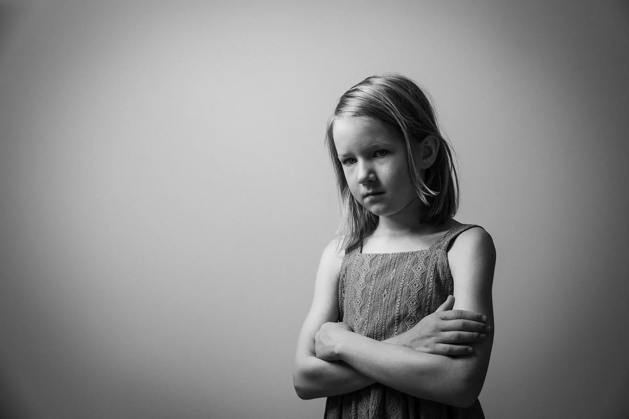 Schwarz-Weiß-Fotografie eines jungen Mädchens mit verschränkten Armen, leicht geneigtem Kopf und ernstem Blick, vor hellem Hintergrund.