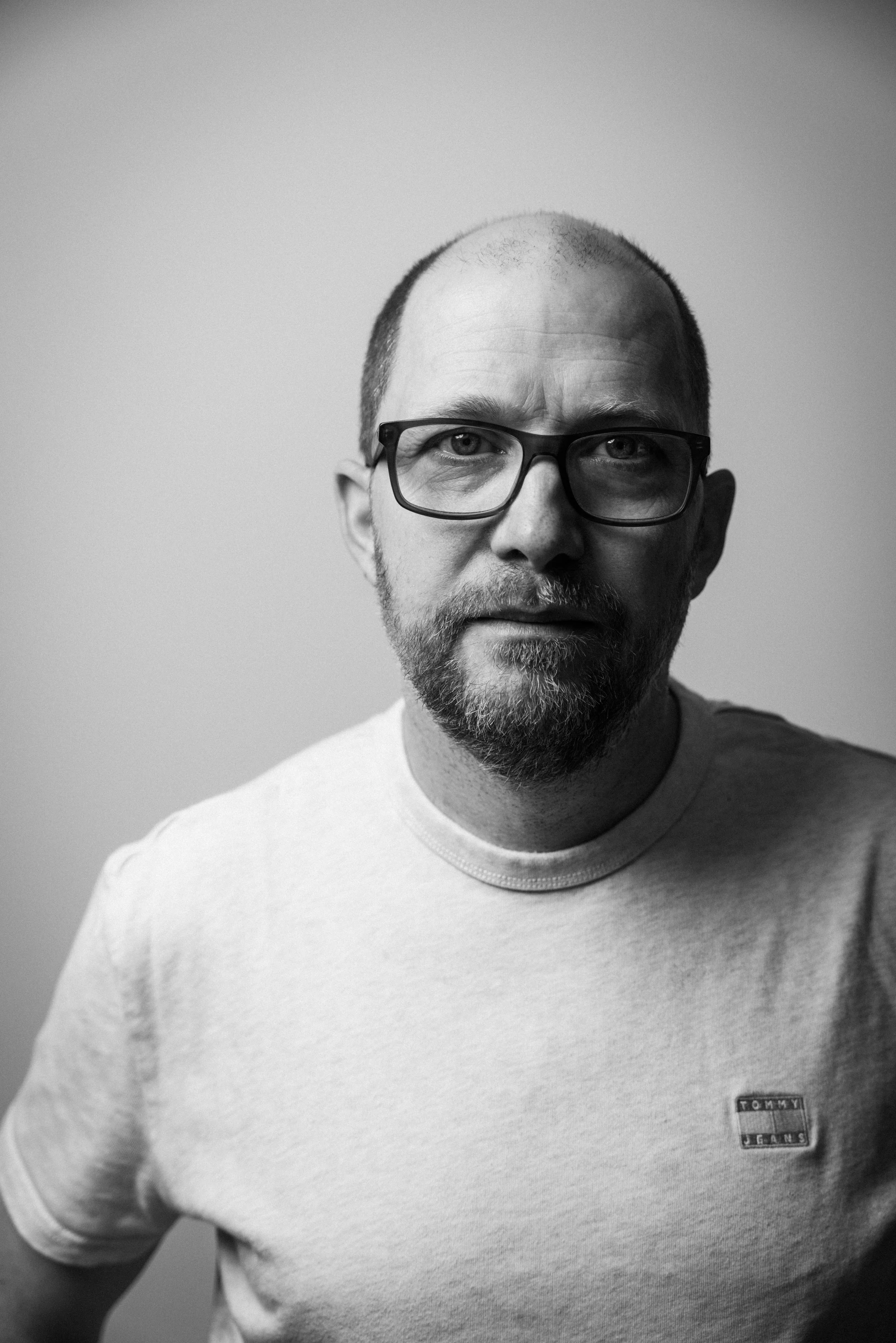 Schwarz-Weiß-Fotografie eines Mannes mit Bart, Brille und kurz geschnittenen Haaren, in einem hellen T-Shirt mit Tommy Hilfiger Logo, vor einem neutralen Hintergrund.
