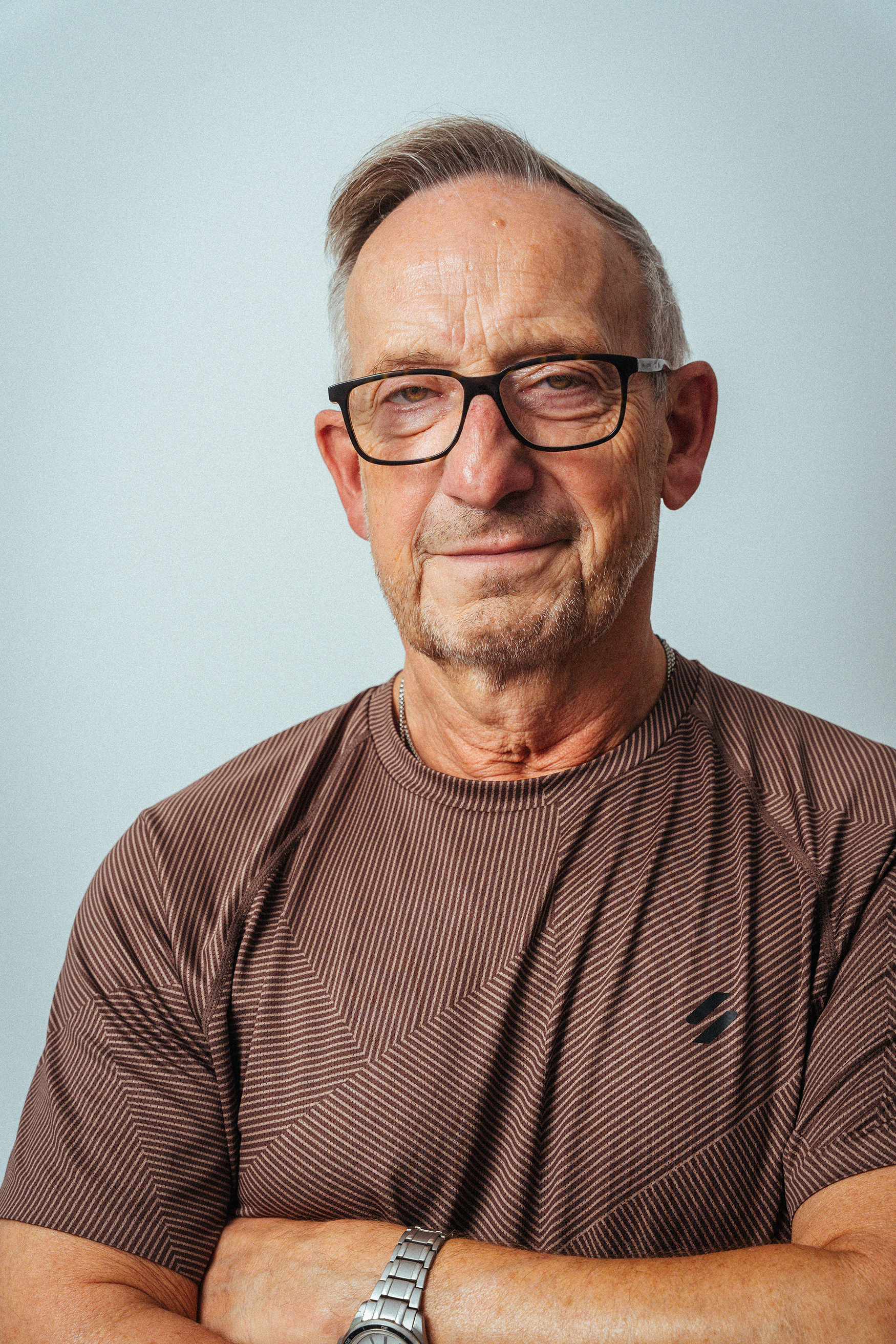 Nahaufnahme eines älteren Mannes mit Brille, kurzen grauen Haaren und einem leichten Lächeln, getragen in einem braunen rippigen T-Shirt. Er steht vor einem hellblauen Hintergrund.