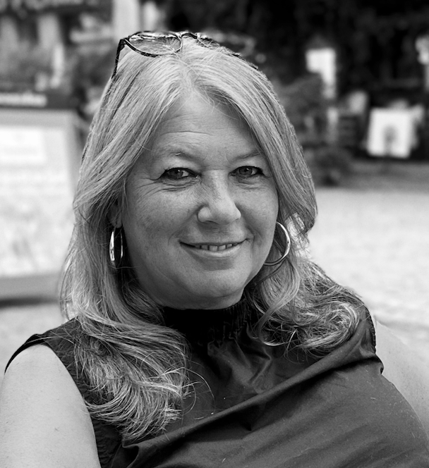 Eine lachende Frau mit langem, lockigem Haar, trägt eine dunkle Bluse und große Ohrringe, im Hintergrund eine Stadt.
