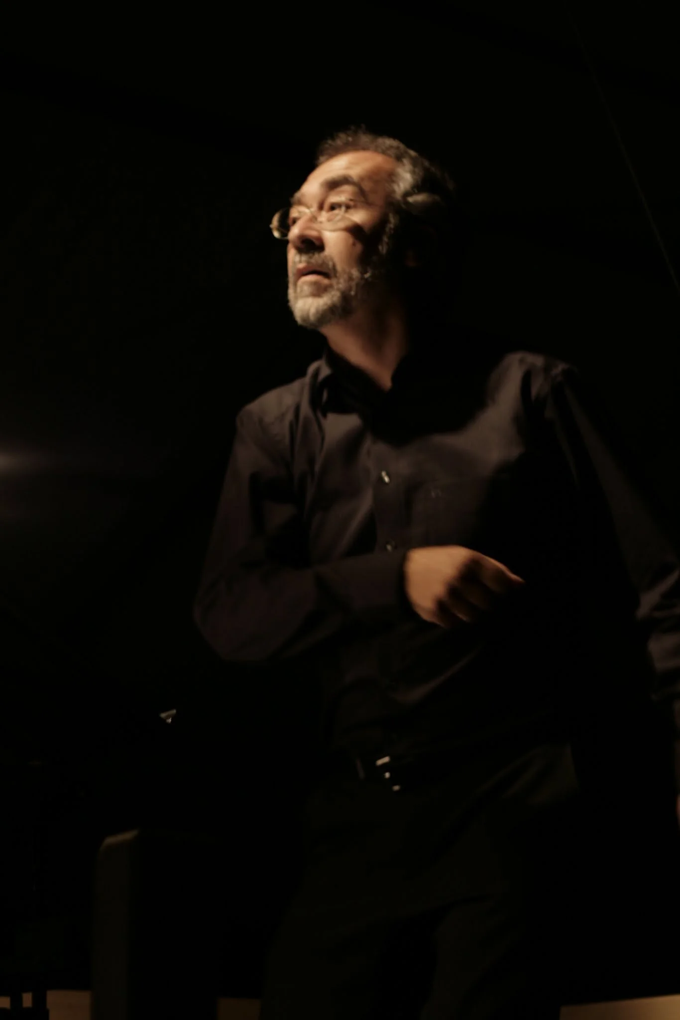 Ein älterer Mann mit Brille, grauem Bart und dunkler Kleidung sitzt in einem dunklen Raum, beleuchtet nur von seitlichem Licht.