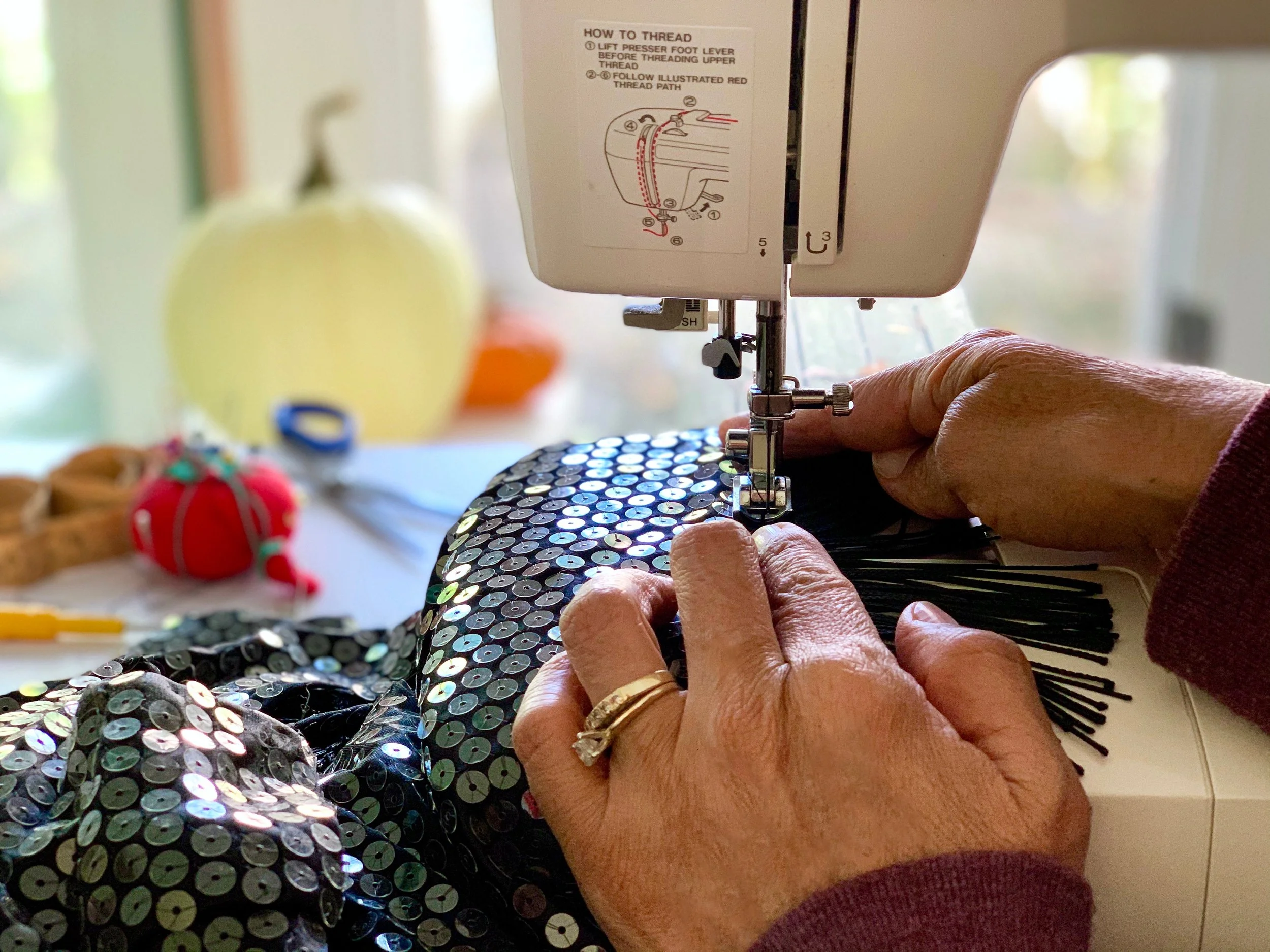 Person näht schwarzes Stoffstück mit schwarzen Pailletten auf Nähmaschine, im Hintergrund Stoffapfel und andere Nähutensilien.