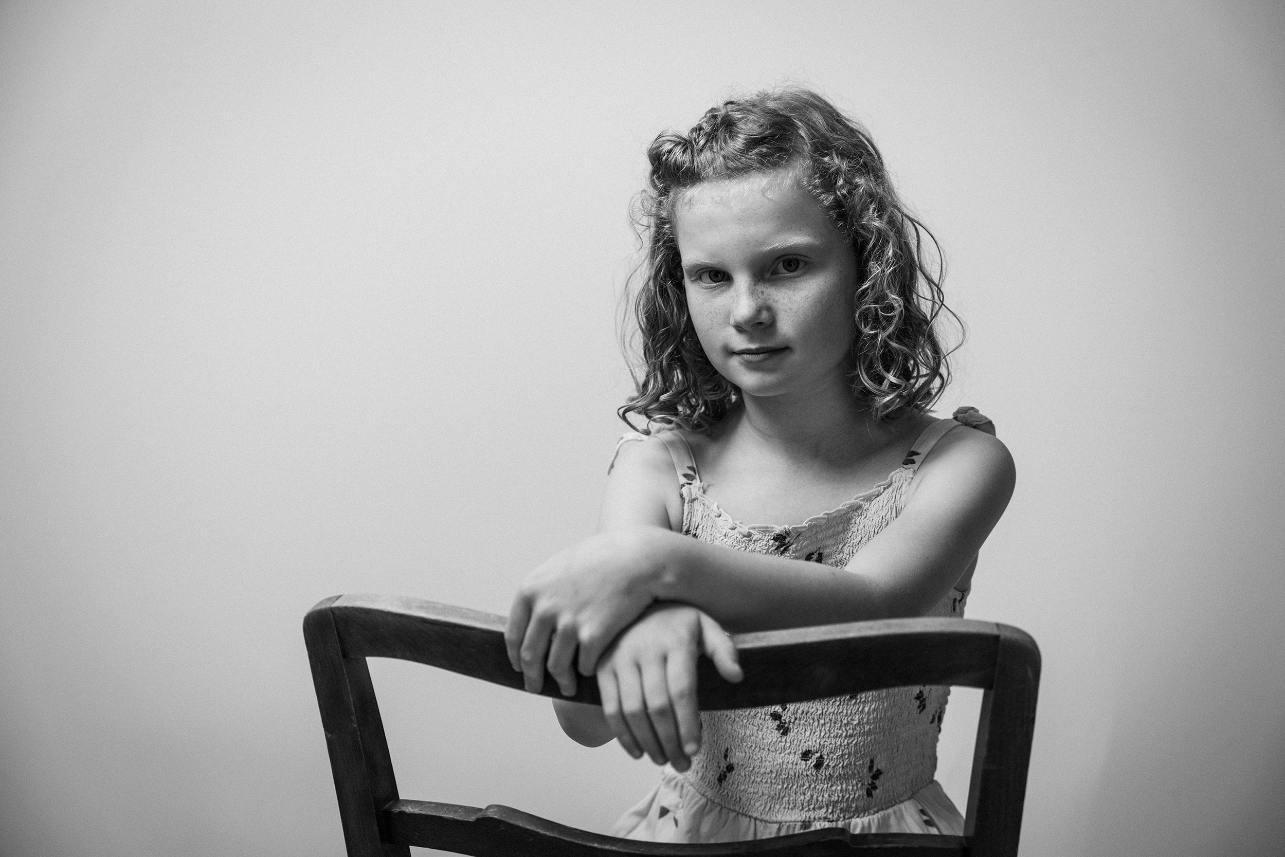 Schwarz-Weiß-Fotografie eines Mädchens, das auf einem Stuhl sitzt, mit verschränkten Armen, vor einem neutralen Hintergrund.