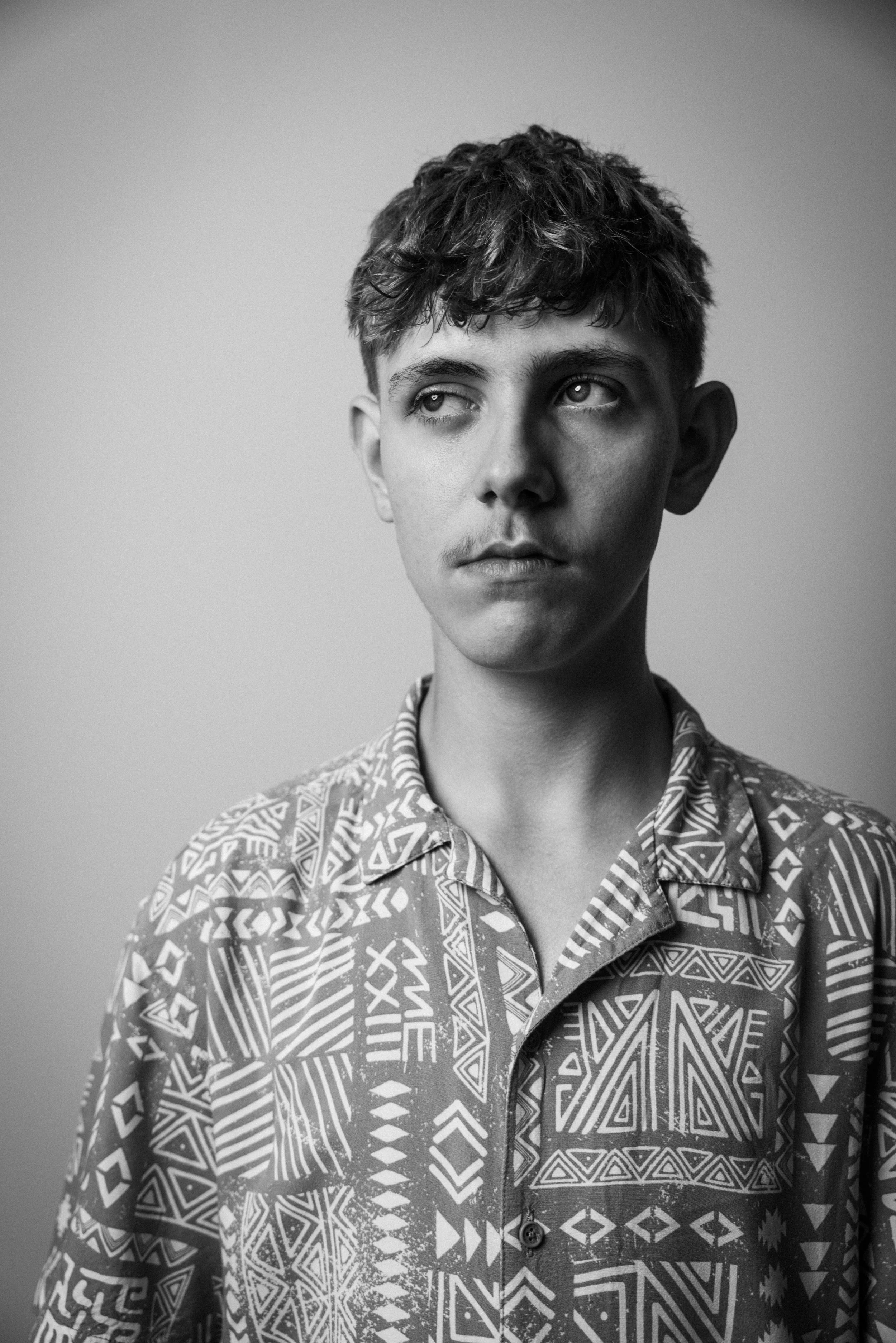 Schwarz-Weiß-Fotografie eines Jungen mit kurzen, gewellten Haaren, der eine bunte, geometrische Muster-Hemden trägt. Der Blick ist nach oben gerichtet, mit nachdenklichem Ausdruck. Der Hintergrund ist neutral und unscharf.