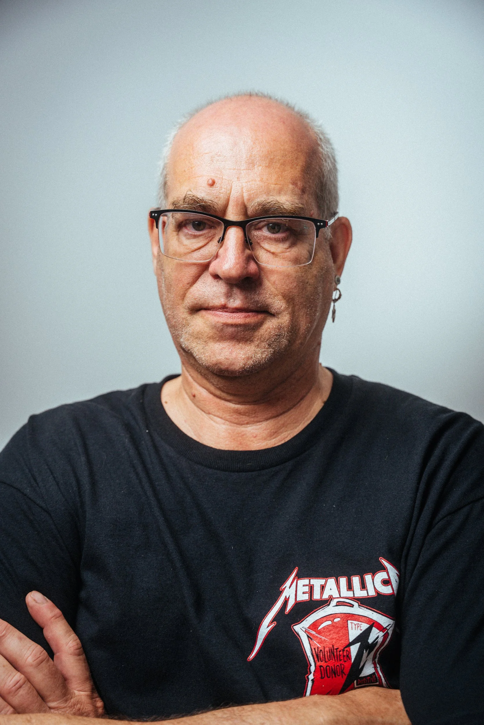 Porträt eines älteren Mannes mit Brille, Earring und weißem Haar, trägt ein schwarzes T-Shirt mit einem Metall-Design und Schriftzug.